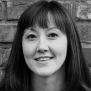 Black and white portrait of a person with long dark hair and bangs, smiling softly. They are posed in front of a brick wall.