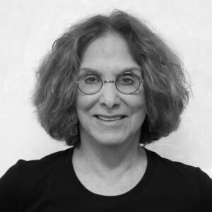 A grayscale photo of a person with shoulder-length curly hair and glasses, smiling while facing the camera. They are wearing a dark shirt against a plain background.