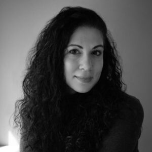 Black and white portrait of a person with long, curly hair, wearing a dark top, looking at the camera with a neutral expression. The background is plain and softly lit.