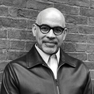 A person with a bald head and a beard, wearing glasses and a leather jacket, stands against a brick wall. The image is in black and white.