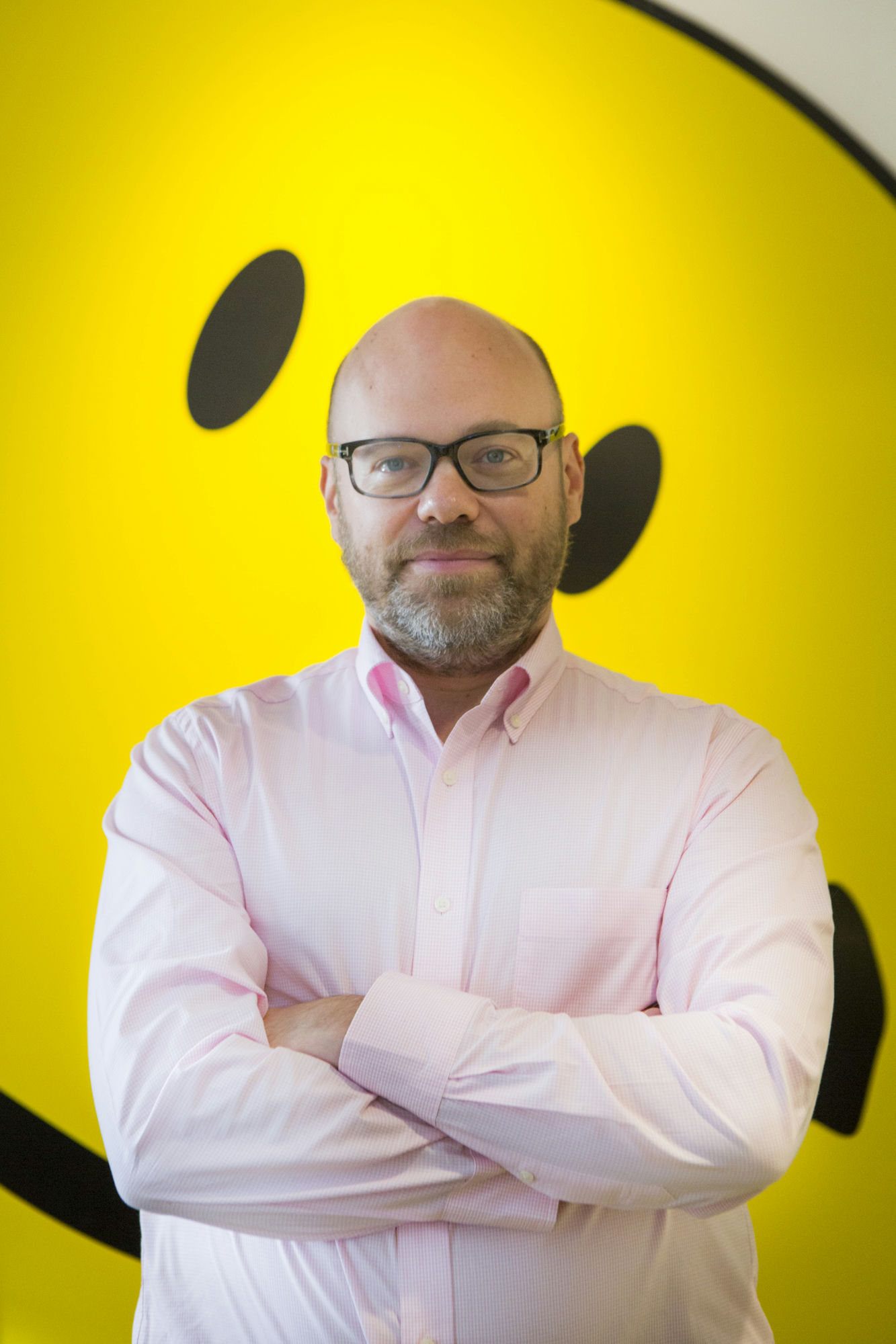 A man with glasses and a beard stands with arms crossed, wearing a light pink shirt. He is in front of a large, bright yellow smiley face background.