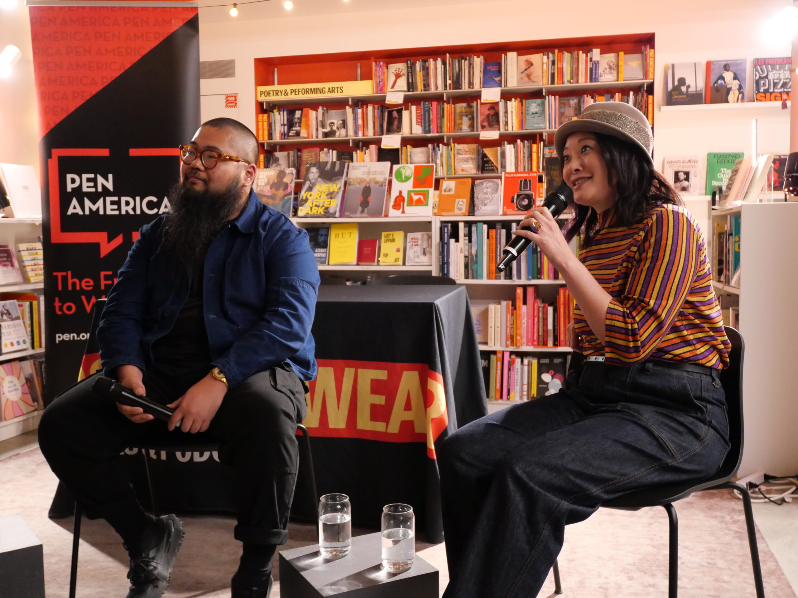 Two people are seated in a bookstore during an event. One holds a microphone, wearing a hat and striped shirt. The other has a notebook, wearing glasses and a blue shirt. A Pen America banner and bookshelves are visible in the background.
