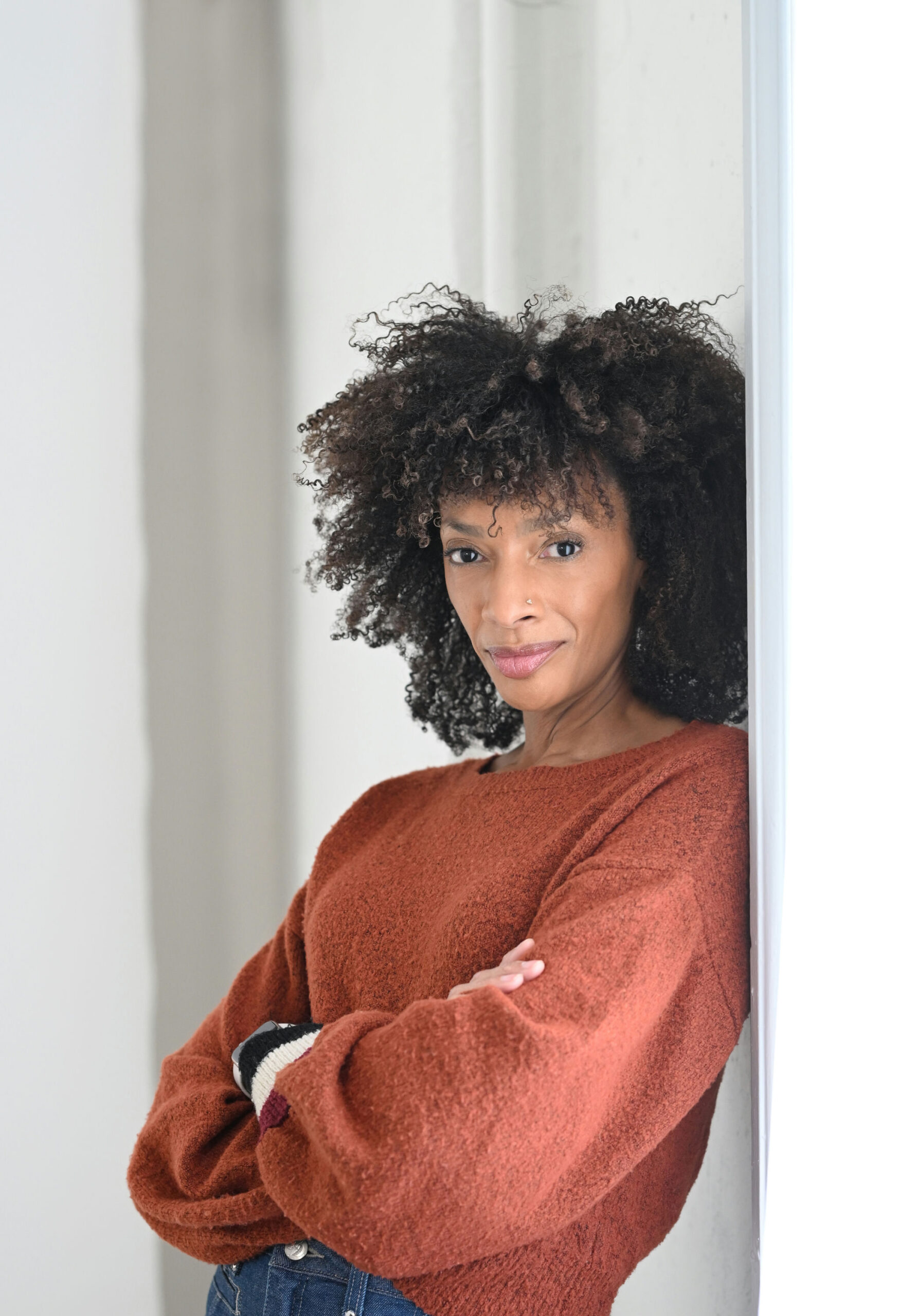 A person with curly hair leans against a light wall, arms crossed. They wear a textured, rust-colored sweater and blue jeans, gazing confidently at the camera with a slight smile, in a softly lit setting.