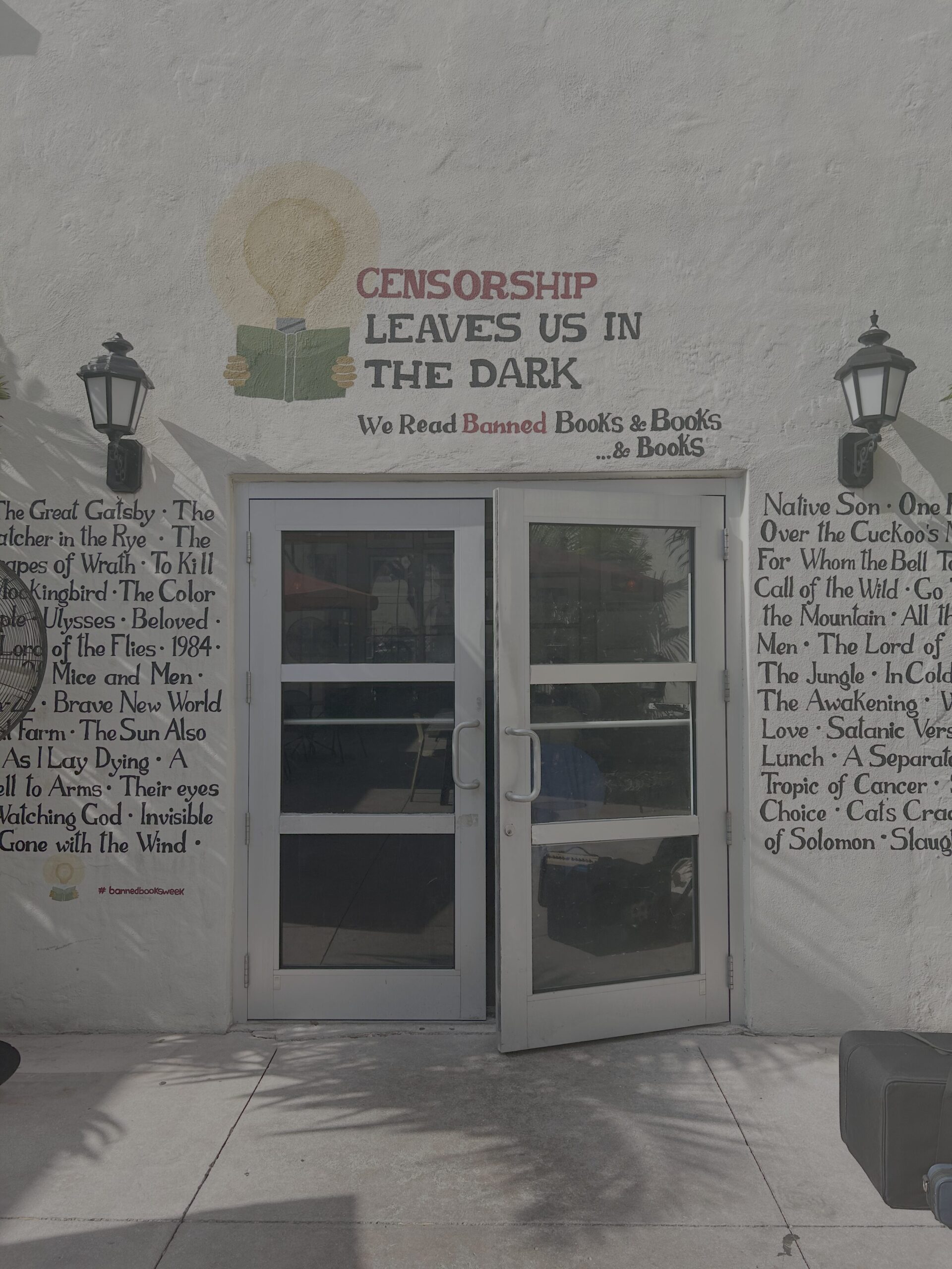 A building wall with the phrase Censorship leaves us in the dark. We read banned books & books... painted above double doors. Titles of various classic books are listed on either side of the doors. Two wall-mounted lanterns are beside the doors.