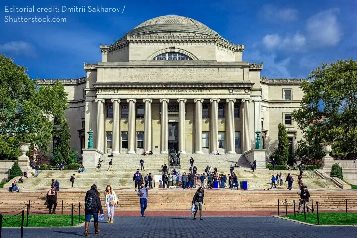 A large neoclassical building with a domed roof and grand columns stands beneath a blue sky. People are sitting and walking on the wide stone steps leading to the entrance. Trees surround the structure.