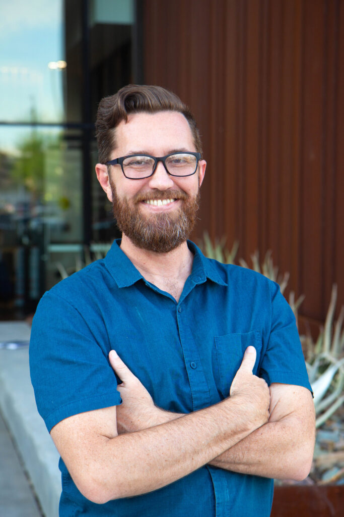 A smiling person with glasses and a beard stands outdoors, with arms crossed, wearing a blue shirt. The background features a modern building with plants in front.