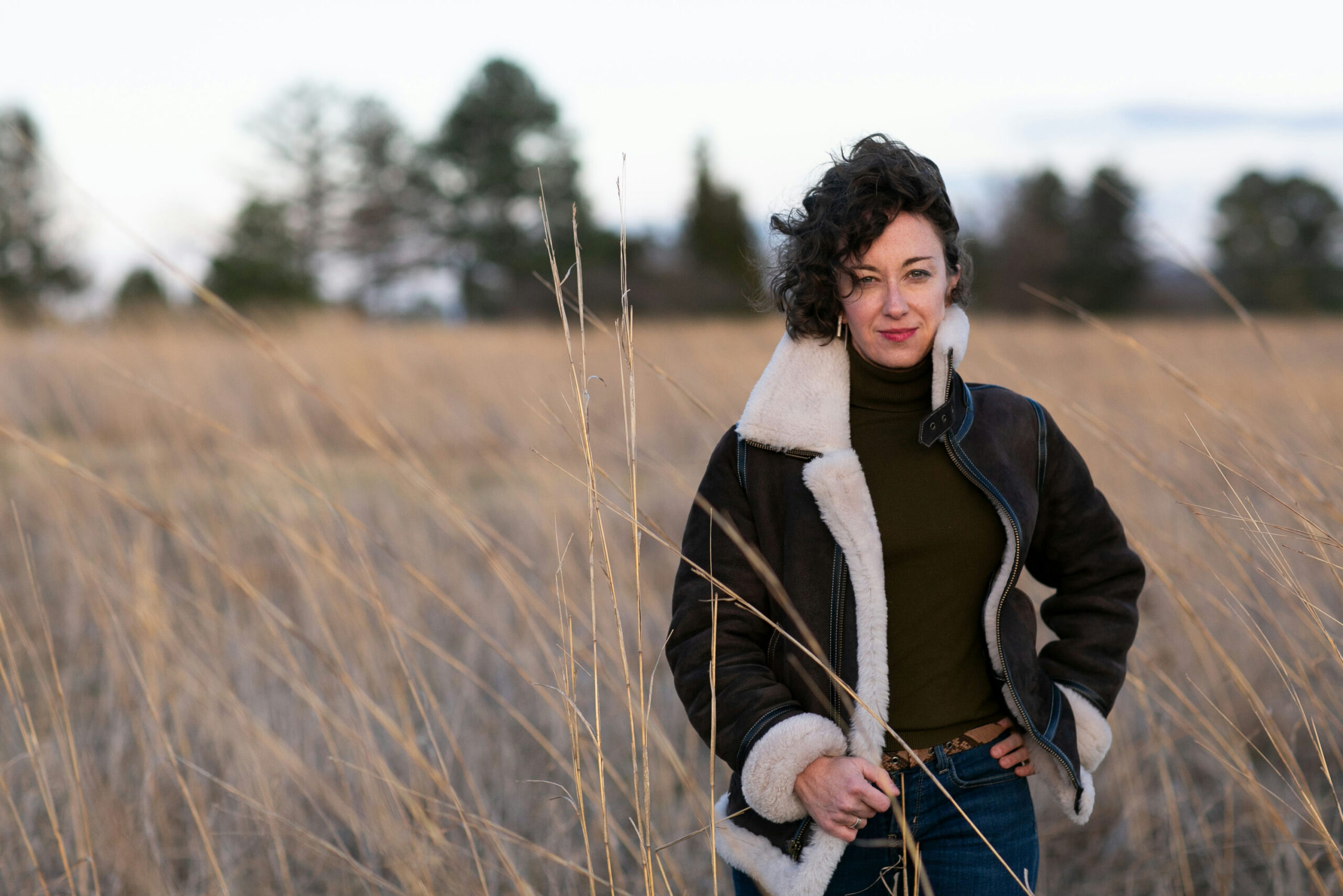 A person with curly hair stands in a field of tall grass, wearing a shearling-lined aviator jacket and jeans. The sky is overcast, and trees are blurred in the background.