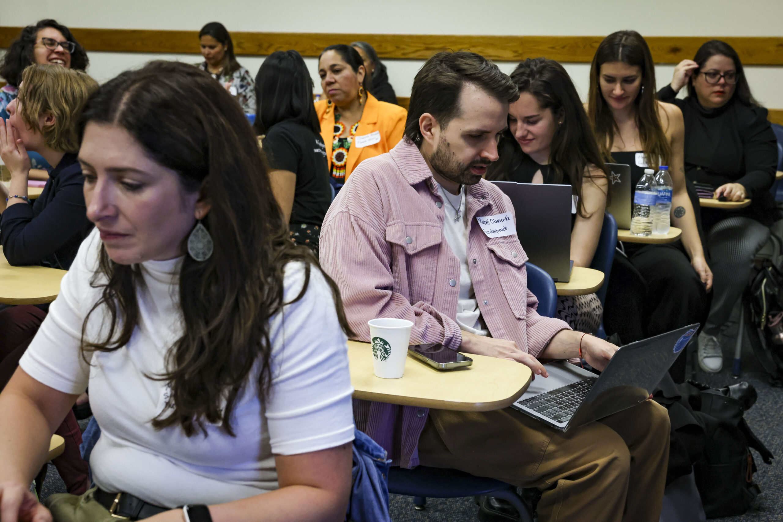 A group of people seated in a classroom, engaging with laptops and notebooks. A man in the center is focused on his laptop, wearing a name tag. The classroom has wooden paneling and rows of desks.