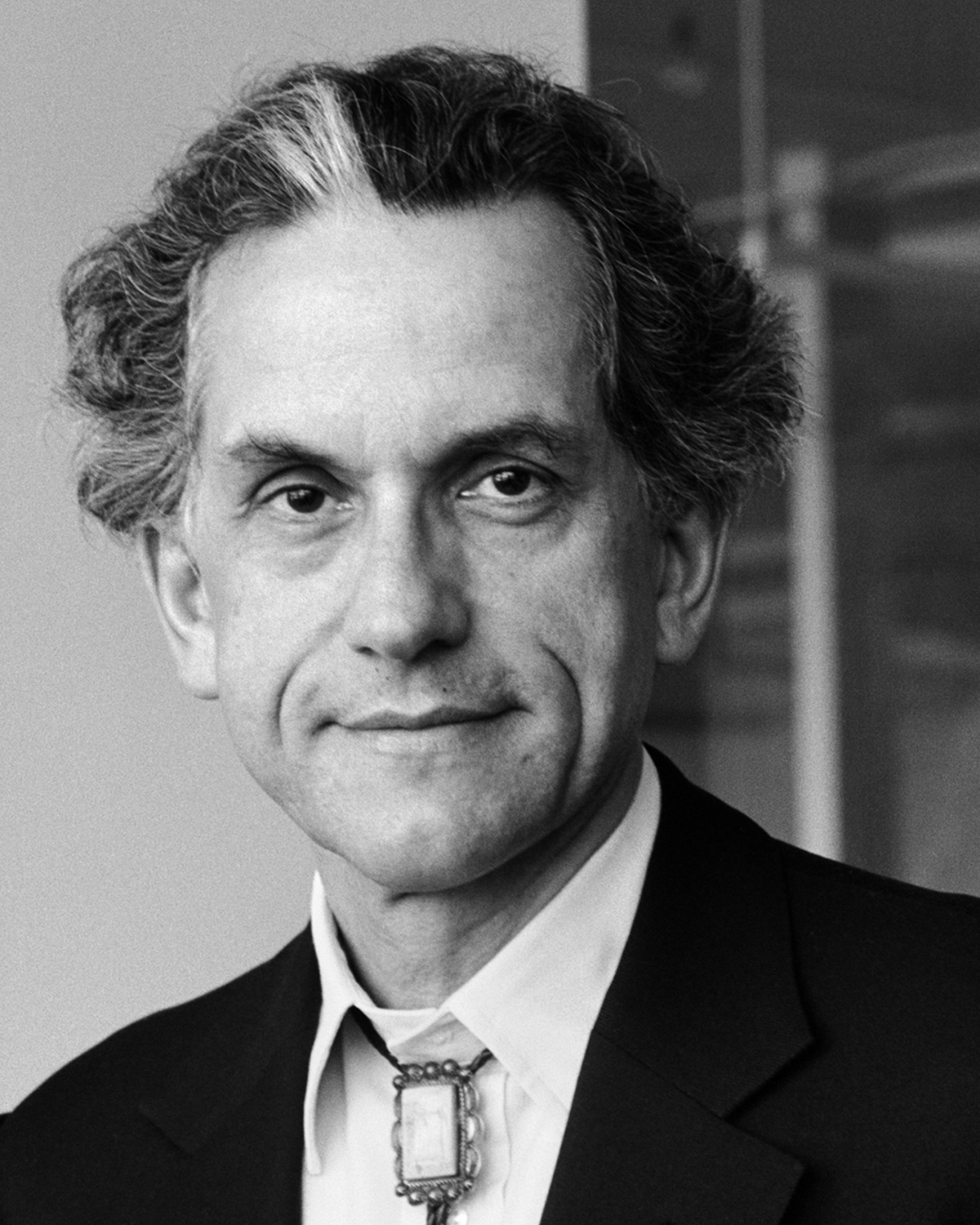 A black and white portrait of a man with wavy hair, wearing a dark suit, white shirt, and bolo tie. He looks directly at the camera with a neutral expression. The background is blurred and indistinct.