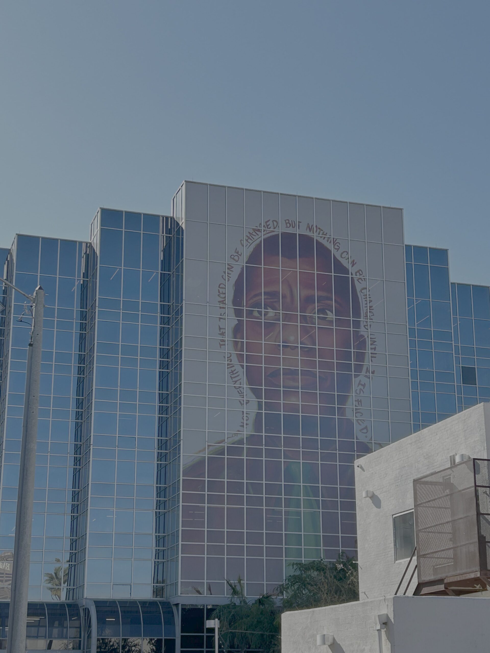 A large mural of a persons face is displayed on the glass facade of a multistory building. The sky is clear, and nearby structures are visible in the foreground. The mural features text surrounding the face.