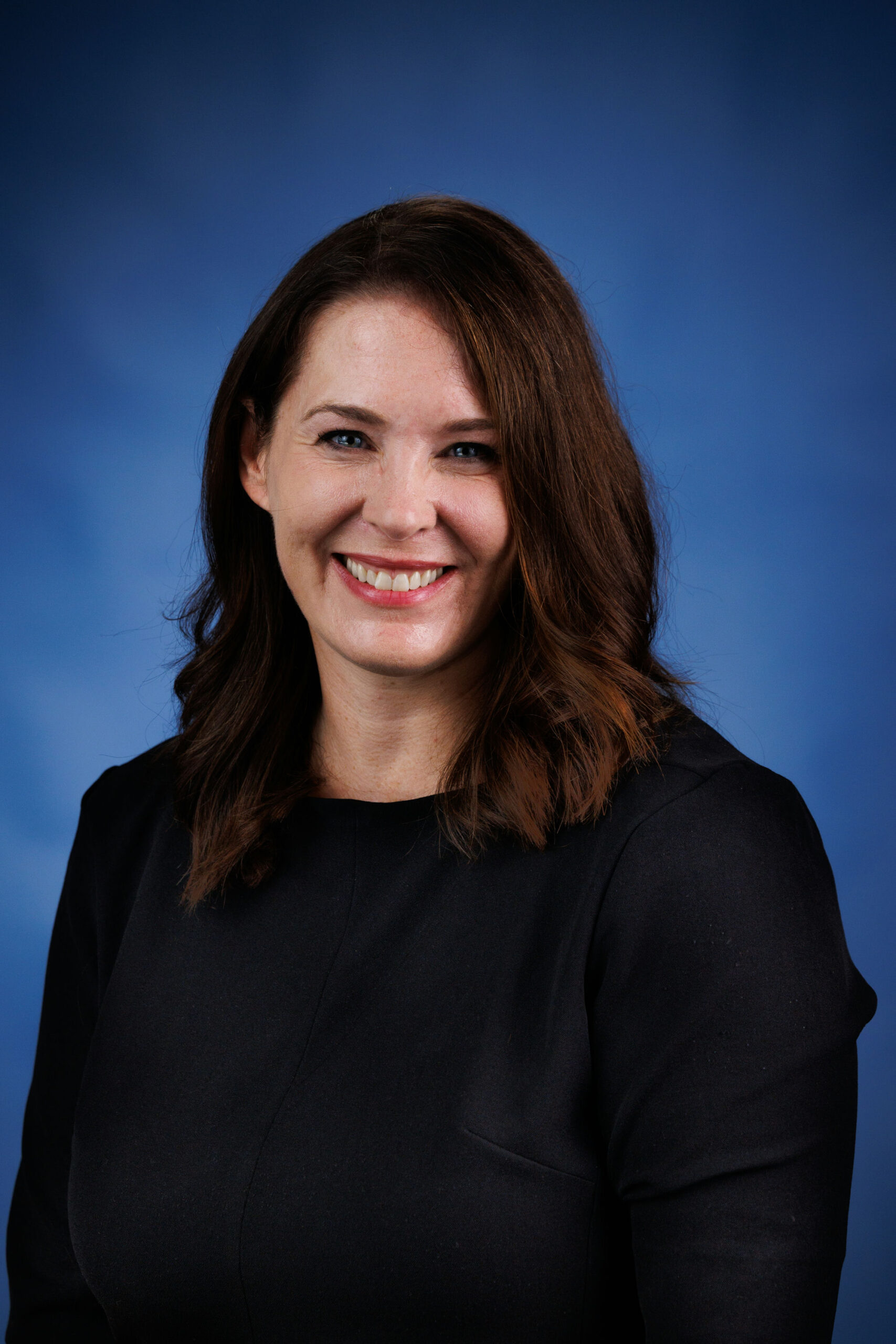 A person with shoulder-length brown hair smiles at the camera against a blue background, wearing a black top.