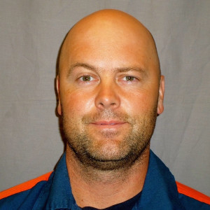 Bald man with light stubble, wearing a blue jacket with orange accents, poses against a plain background.