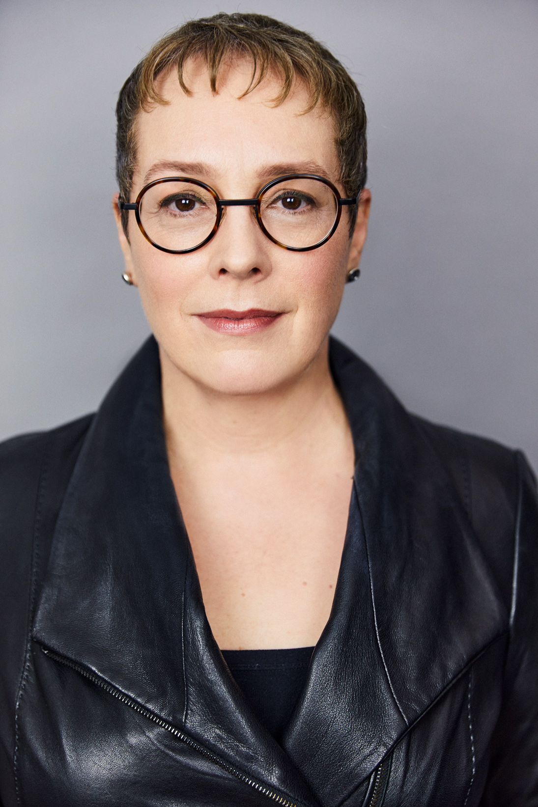 A person with short hair, wearing round glasses and a black leather jacket, stands against a gray background. Their expression is neutral.
