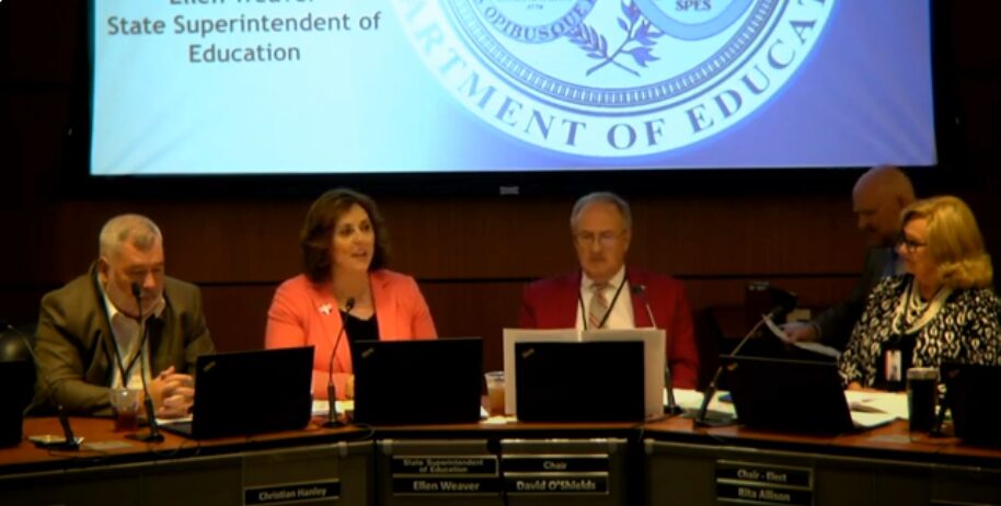 Five people sit at a conference table with microphones. A woman in a pink blazer speaks, while the others listen. A large screen behind them displays the Department of Education logo.