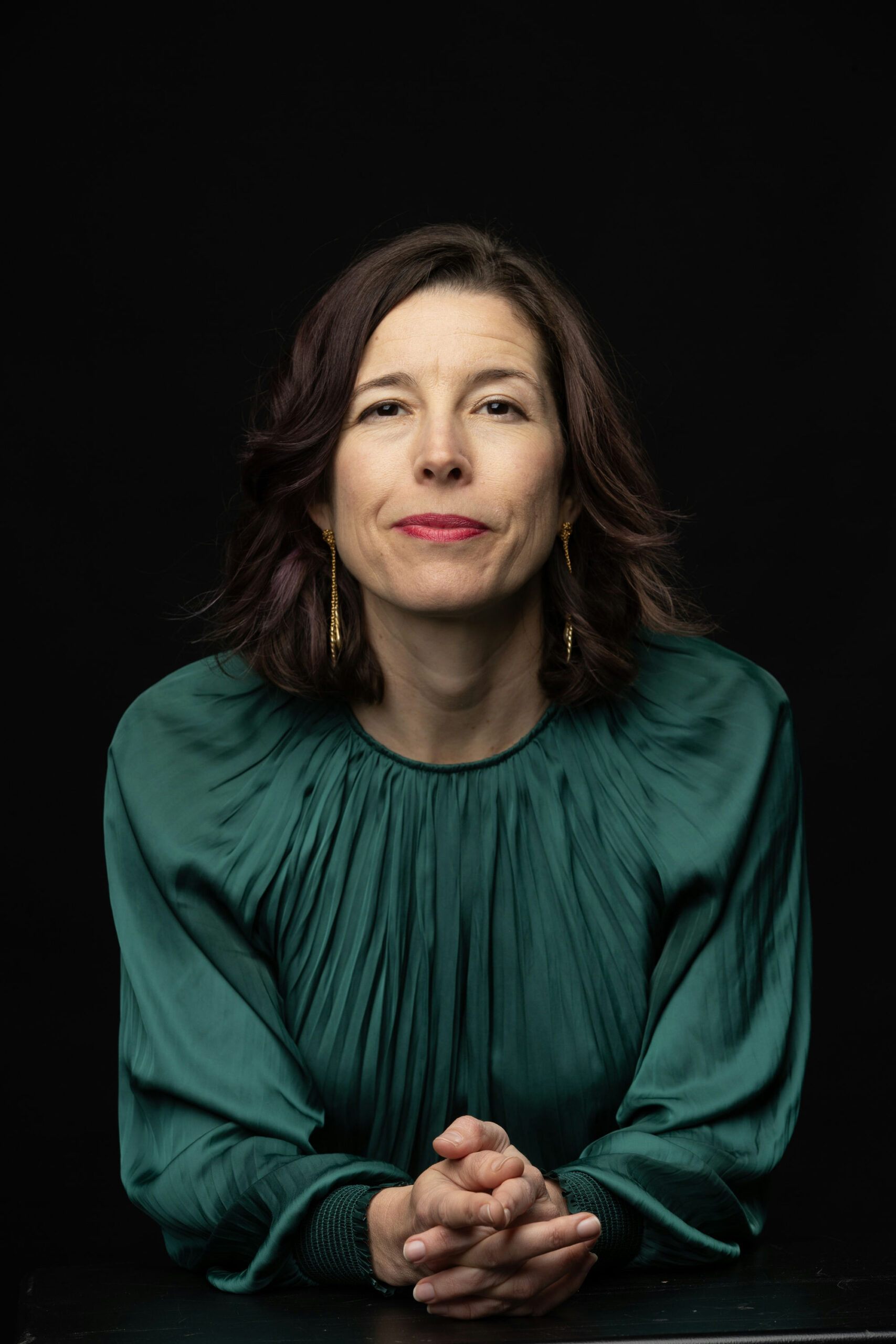 A woman with shoulder-length brown hair is wearing a green blouse. She is seated against a black background, facing the camera, with her hands clasped in front of her and a neutral expression.