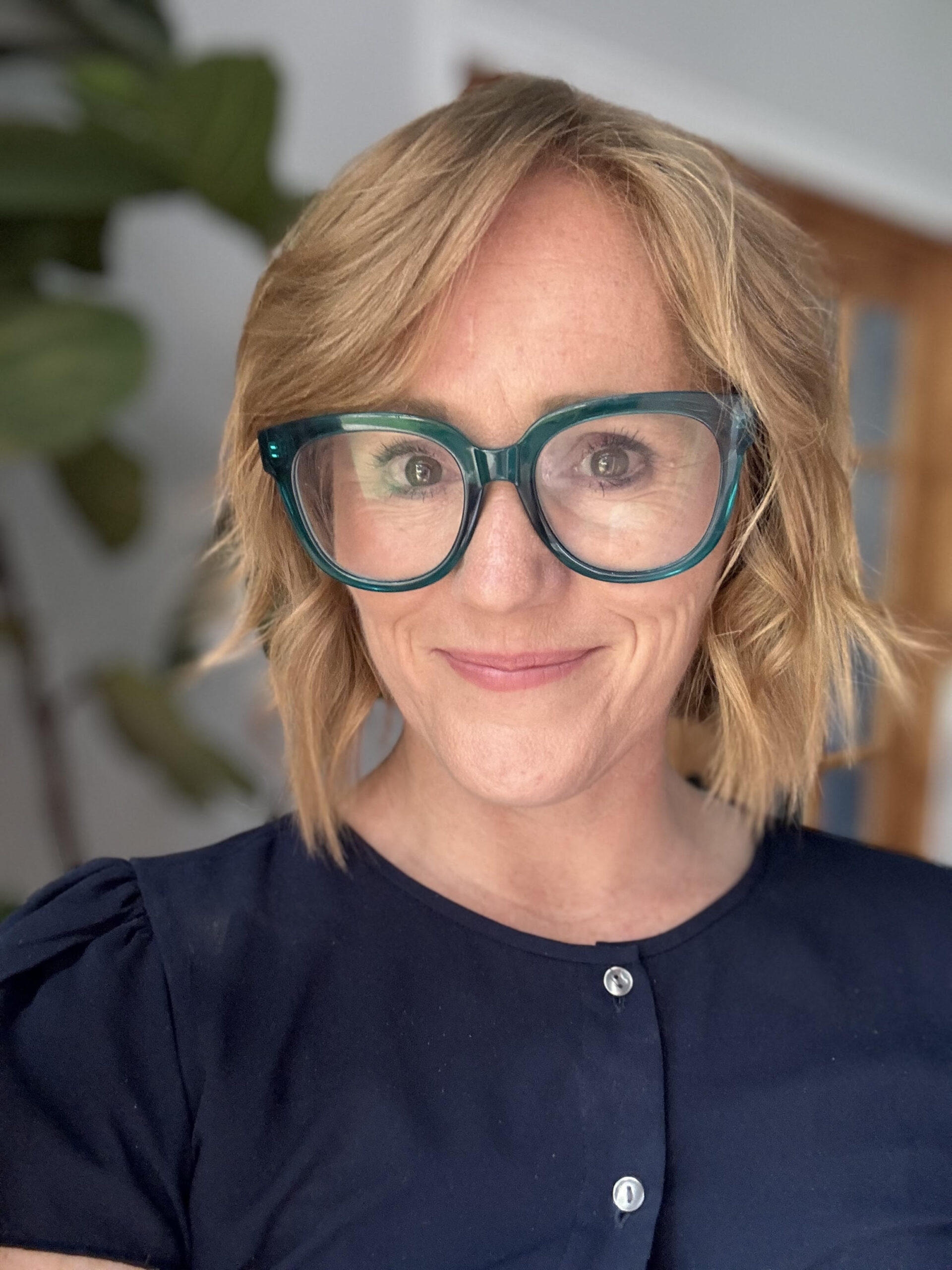 A person with short, wavy blonde hair is smiling at the camera. They are wearing oversized teal glasses and a navy blue top with buttons. Theres a blurred plant in the background.