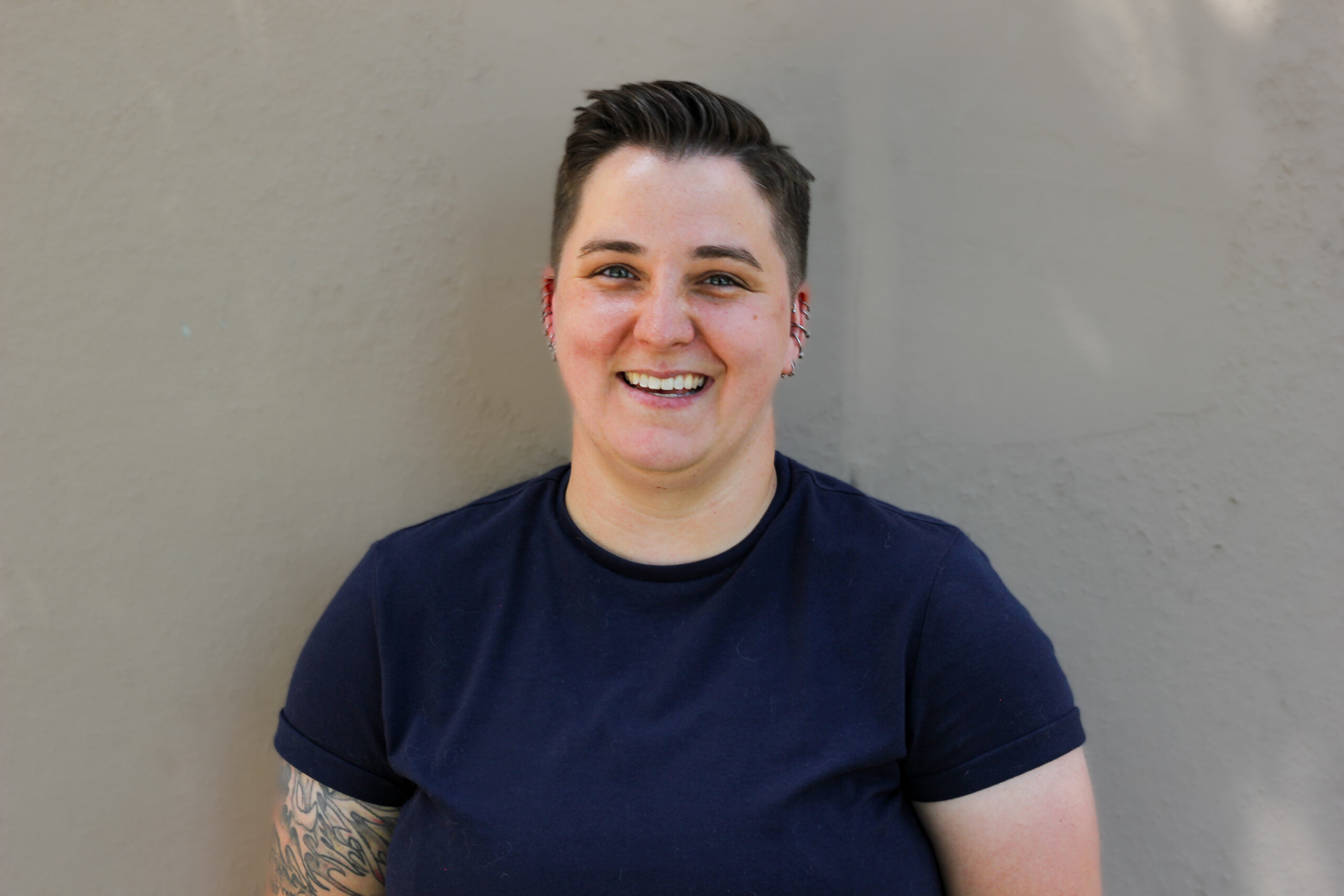 A person with short hair is smiling while standing against a light-colored wall. They are wearing a dark T-shirt and have visible tattoos on one arm.