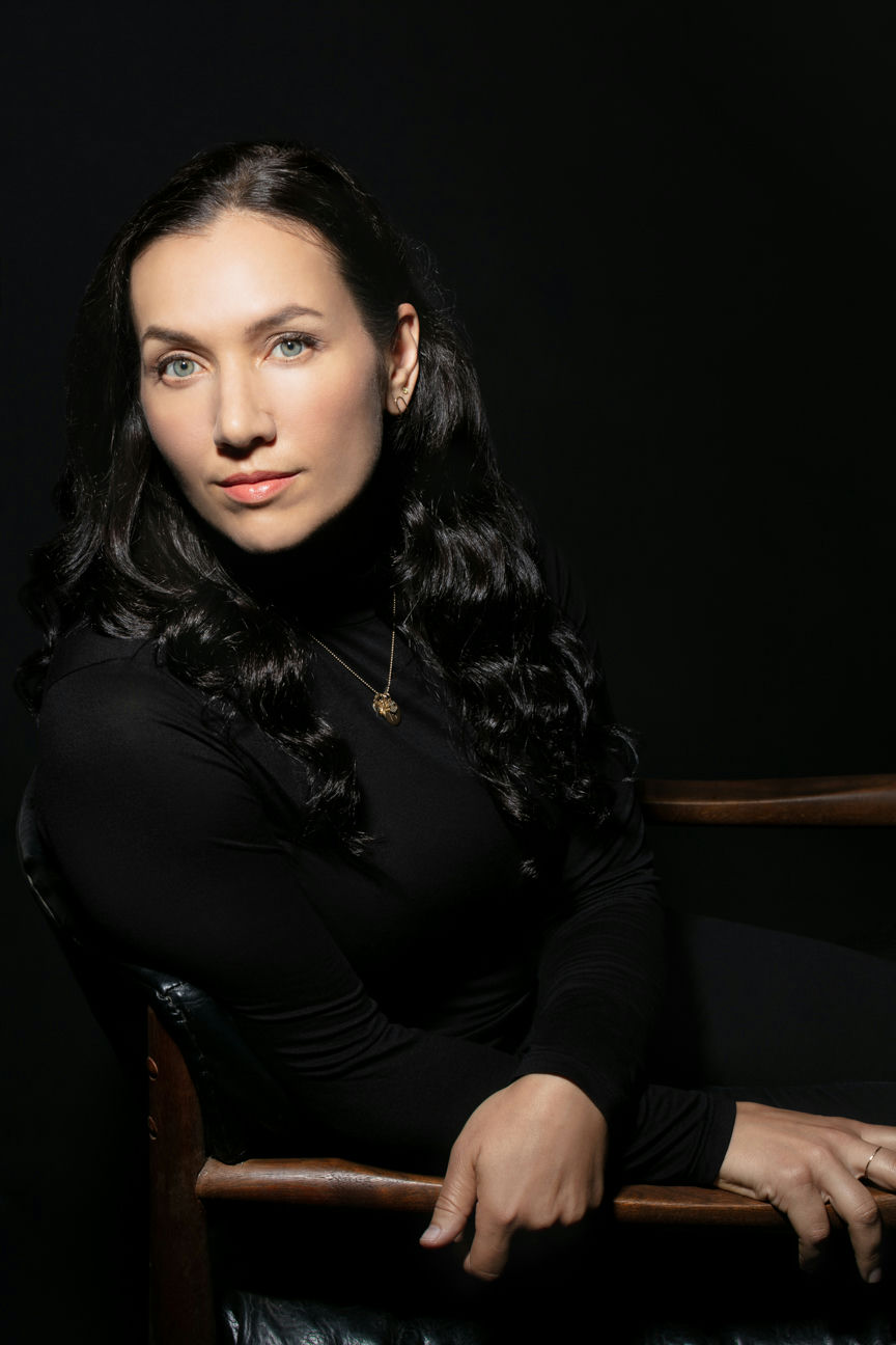 A woman with long, dark hair and a black turtleneck sits in a wooden chair against a dark background. Her hands rest on the chairs arm while she gazes directly at the camera, wearing a subtle necklace.