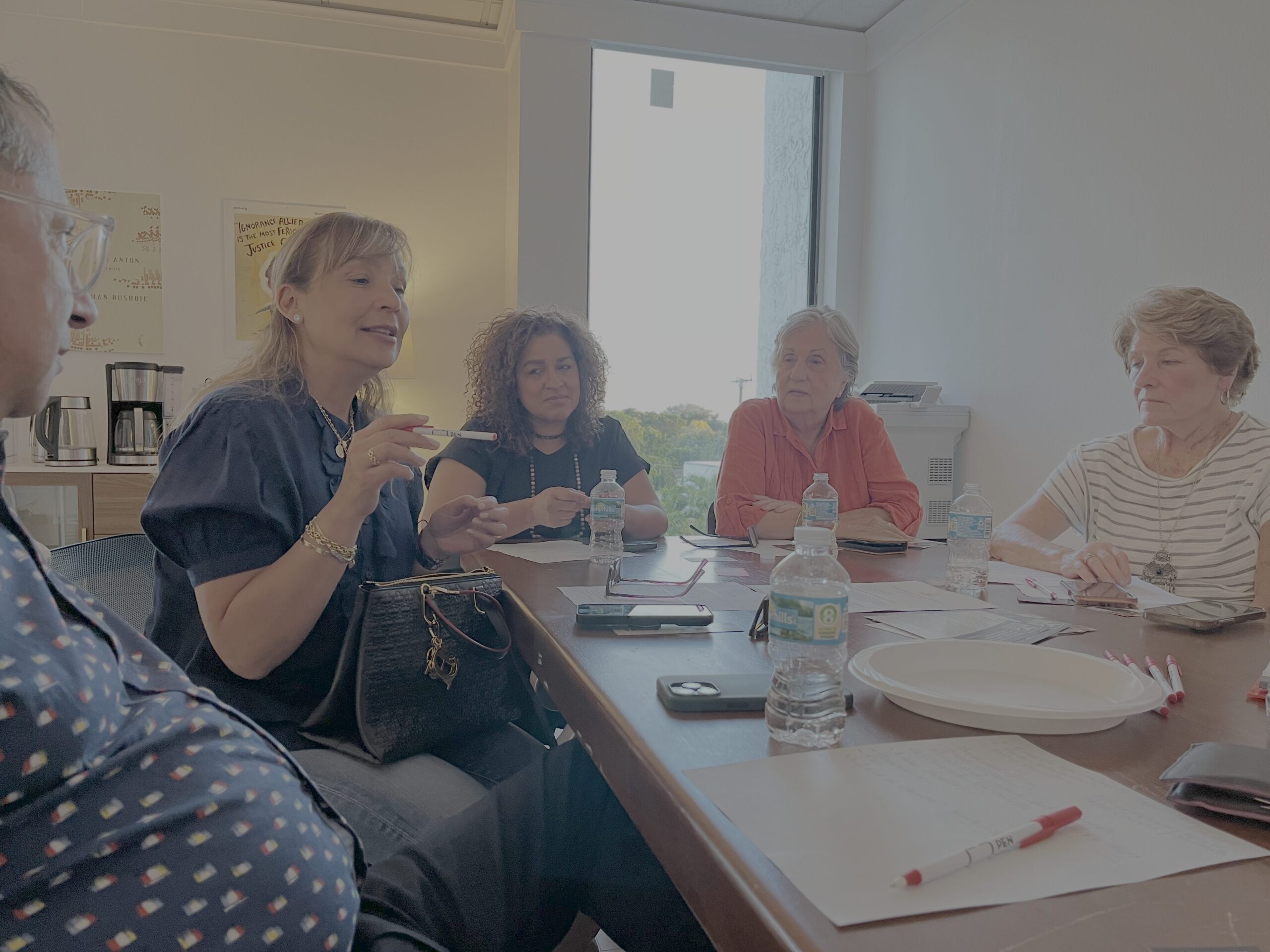 A group of five people sitting around a table having a discussion. The table has notebooks, smartphones, bottled water, and a plate. They are in a well-lit room with a large window in the background.