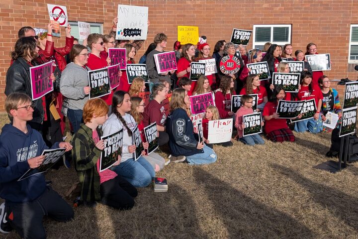 A group of people, mostly teens, kneels outside holding protest signs advocating for freedom to read. They display messages like Freedom to Read and Read Banned Books. A brick building is visible in the background.