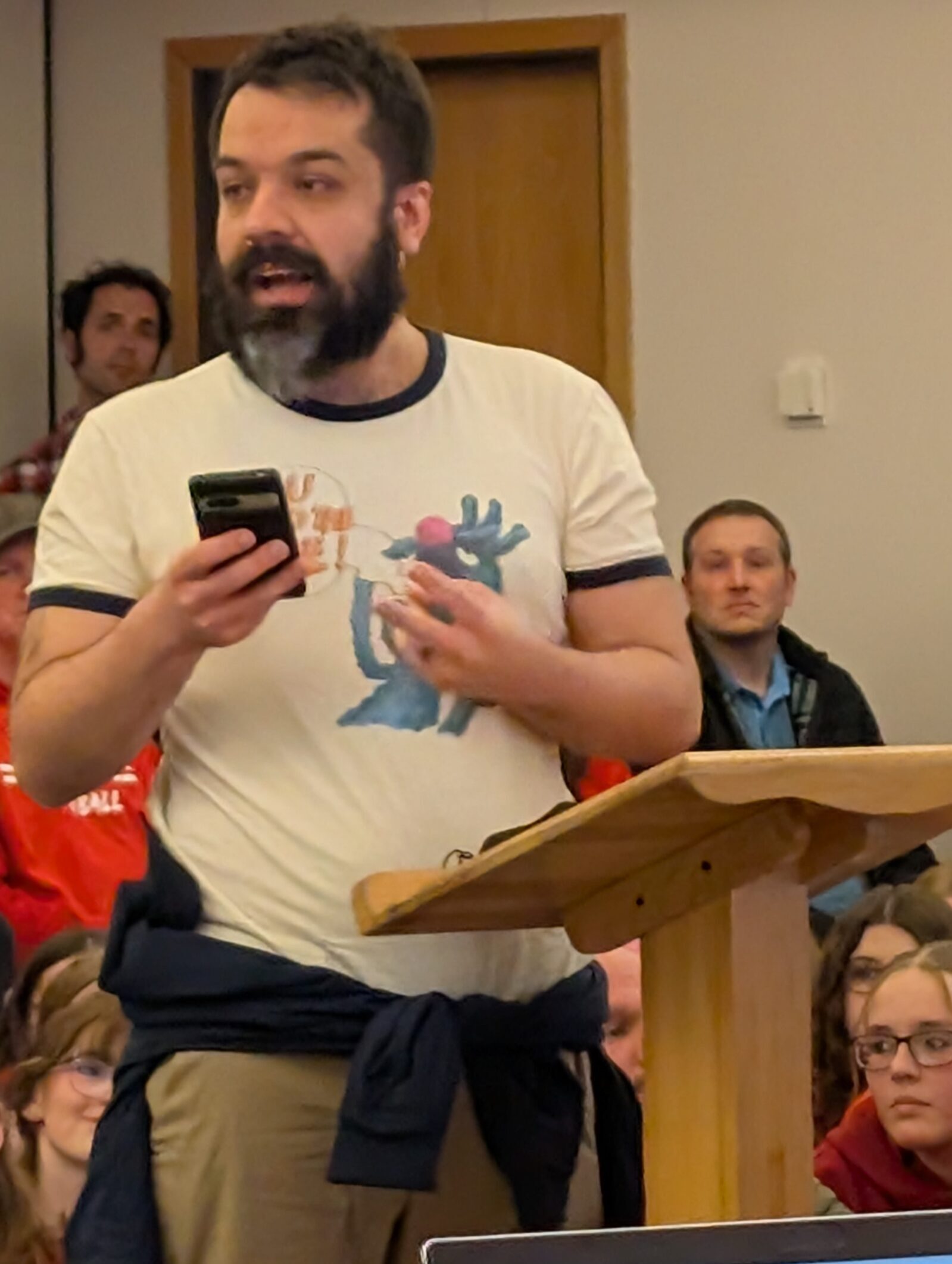 A bearded man speaks at a wooden podium, holding a phone. Wearing a graphic T-shirt and with a jacket tied around his waist, he addresses the issue of St. Francis book bans. Seated people, some in red shirts, watch attentively while a laptop with an open document is visible in the foreground.