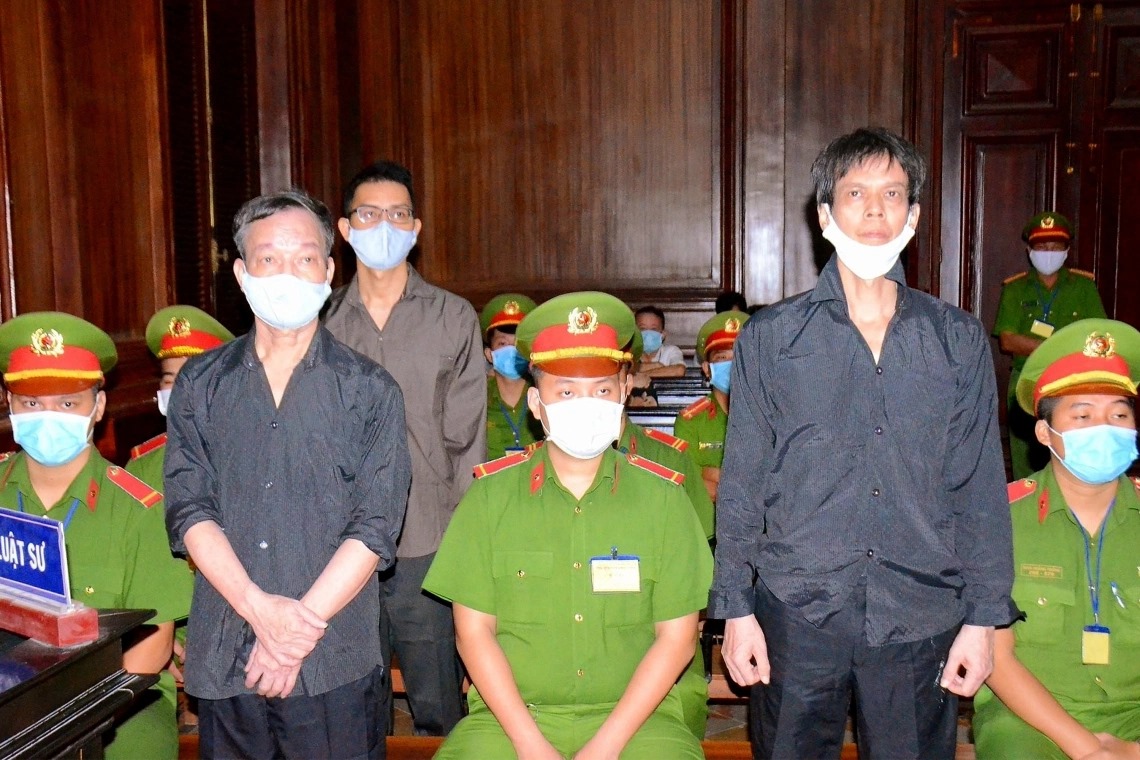 A group of people standing in what appears to be a courtroom. Two men in black shirts stand in front, while several individuals in green uniforms with badges are seated and standing behind them. All are wearing face masks.