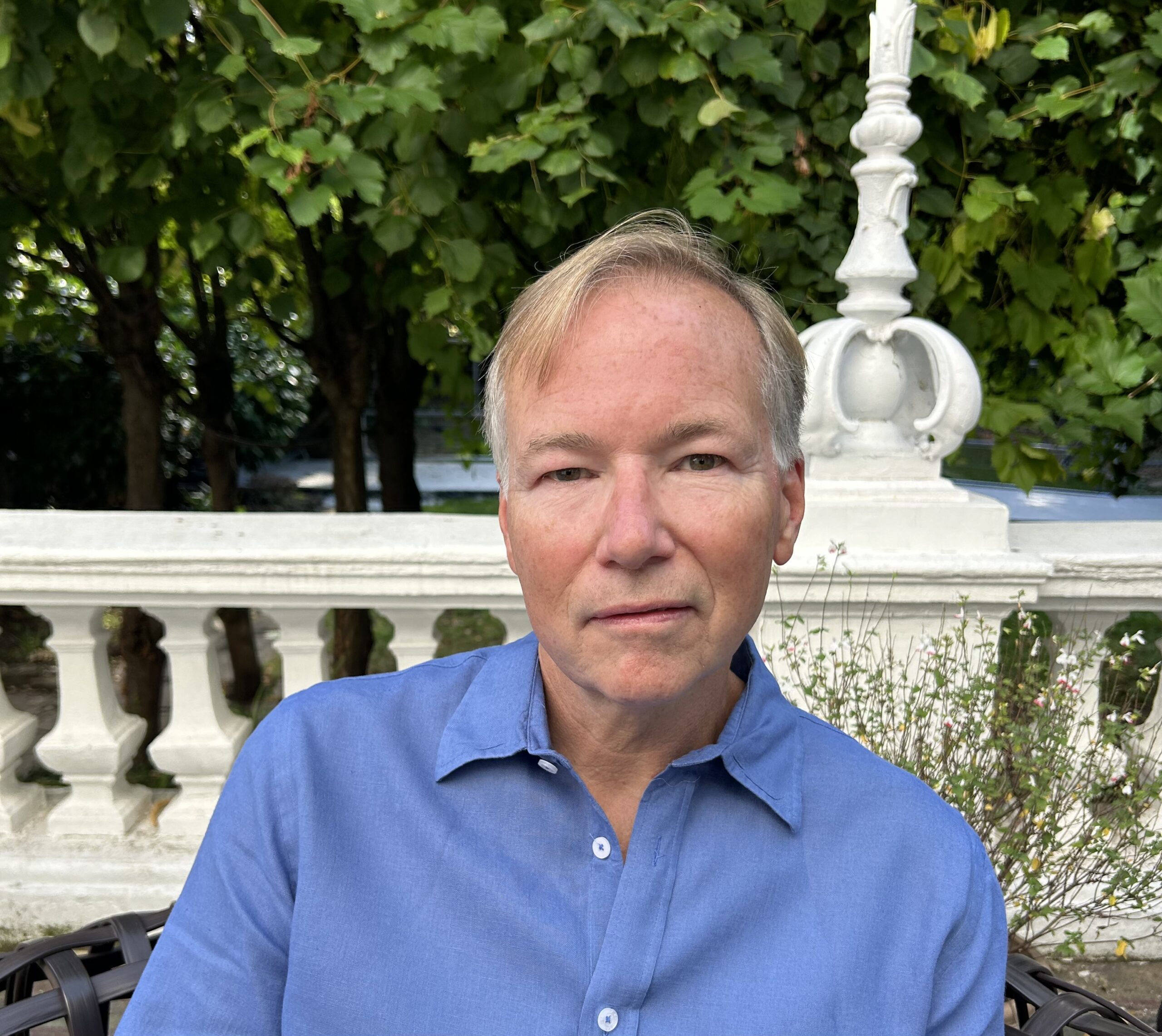 A person with light hair is sitting outdoors in front of a white stone railing and lush greenery. They are wearing a blue shirt and looking at the camera with a neutral expression.
