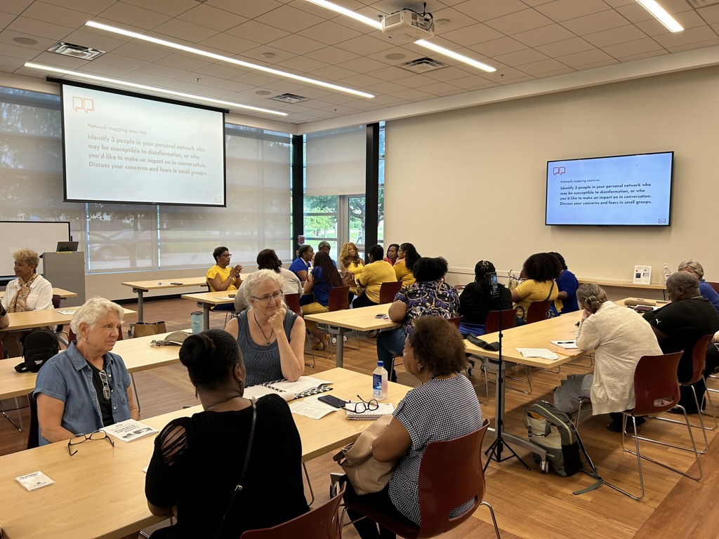 A group of people with name tags seated at tables in a well-lit room, engaged in conversation. Two large screens on the walls display instructions for discussion groups. Many attendees are in blue and yellow shirts.