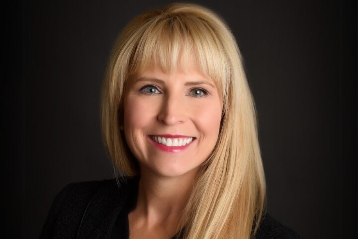 A woman with long blonde hair smiles warmly. She is wearing a black top, and the background is dark, making her hair and smile stand out. Her expression is friendly and approachable.