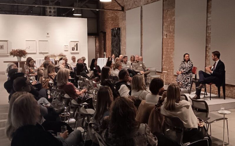 A group of people seated in a room with exposed brick walls, listening to a woman and a man speaking in front. The room is softly lit, with art pieces hanging on white walls in the background.