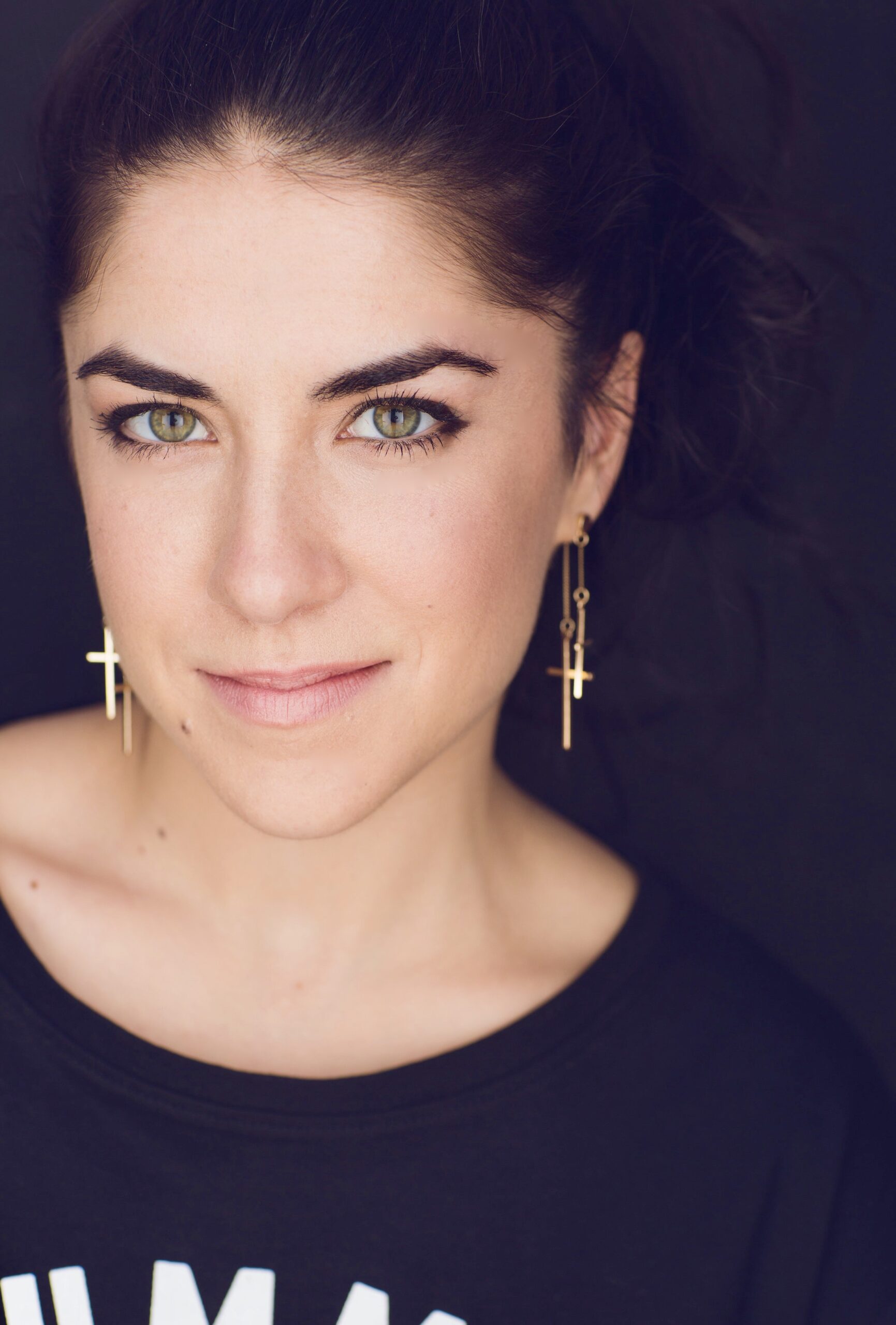 A woman with light eyes, dark hair tied back, and gold cross earrings smiles softly at the camera against a dark background. She wears a black top and subtle makeup.