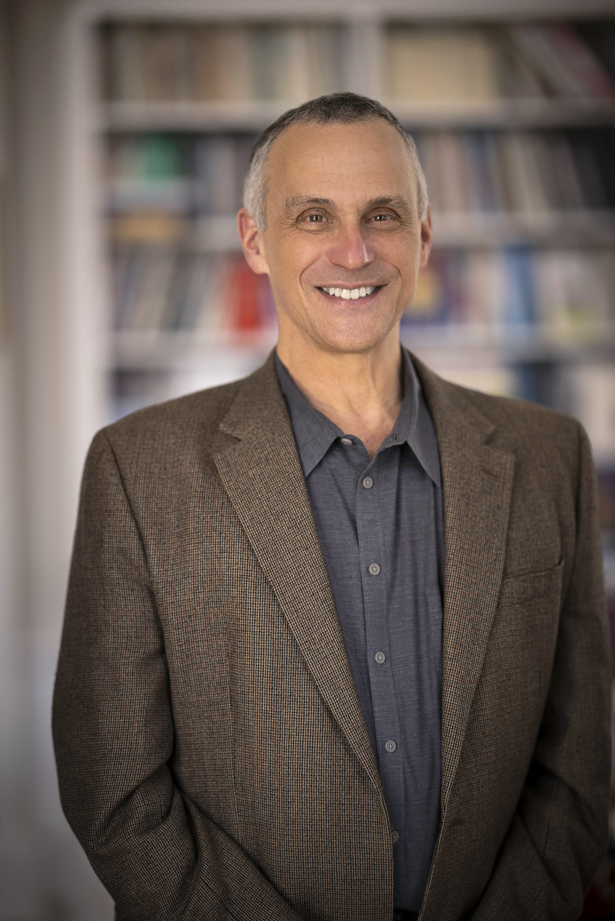 A smiling man in a brown blazer and gray shirt stands in front of blurred bookshelves.