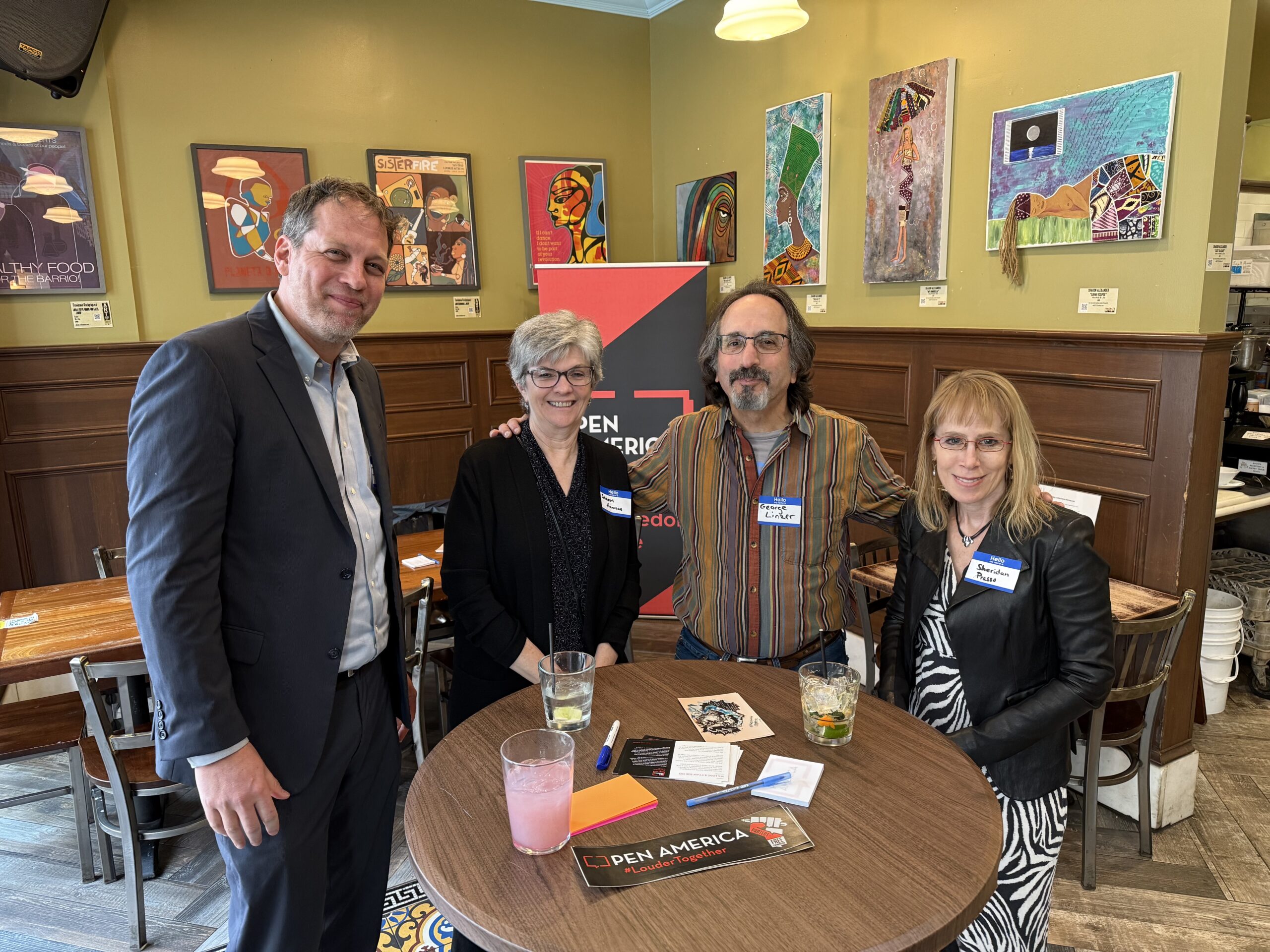 Four people stand and smile around a round table at an indoor event with colorful art on the walls. There are drinks on the table, and a PEN America sign is visible in the background.
