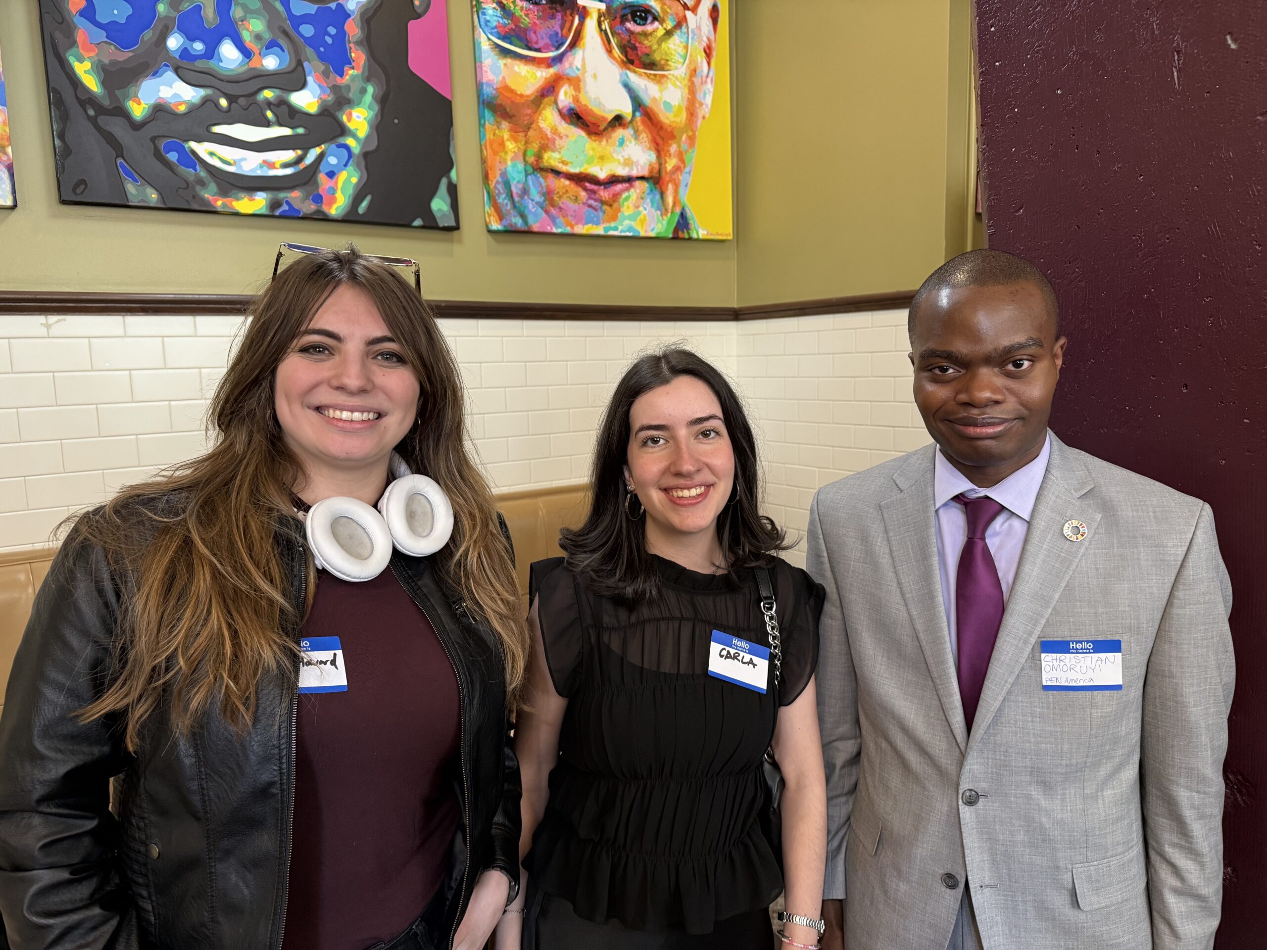 Three people stand together indoors, smiling at the camera. Two women on the left and a man in a suit on the right, all wearing name tags. Colorful artwork is visible on the wall behind them.