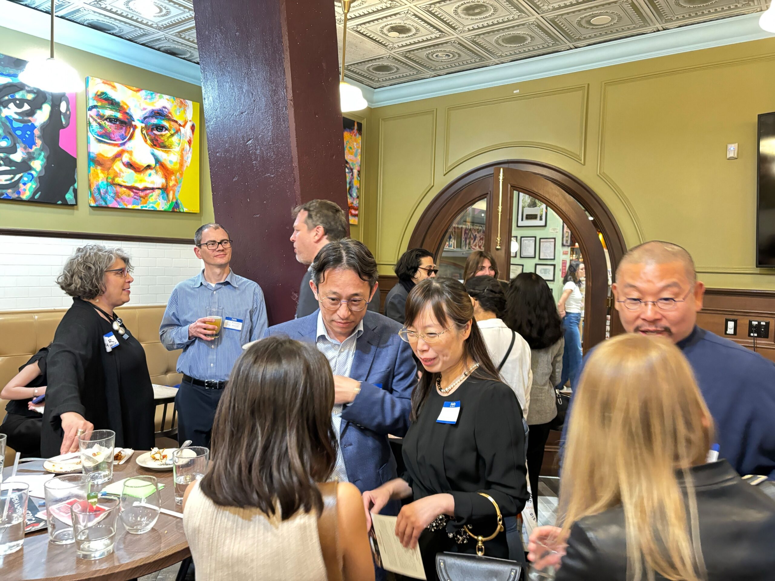 A group of people socializing in a restaurant with drinks and snacks on a table. The room has colorful artwork on the walls and an ornate ceiling. People are wearing name tags and seem engaged in conversation.