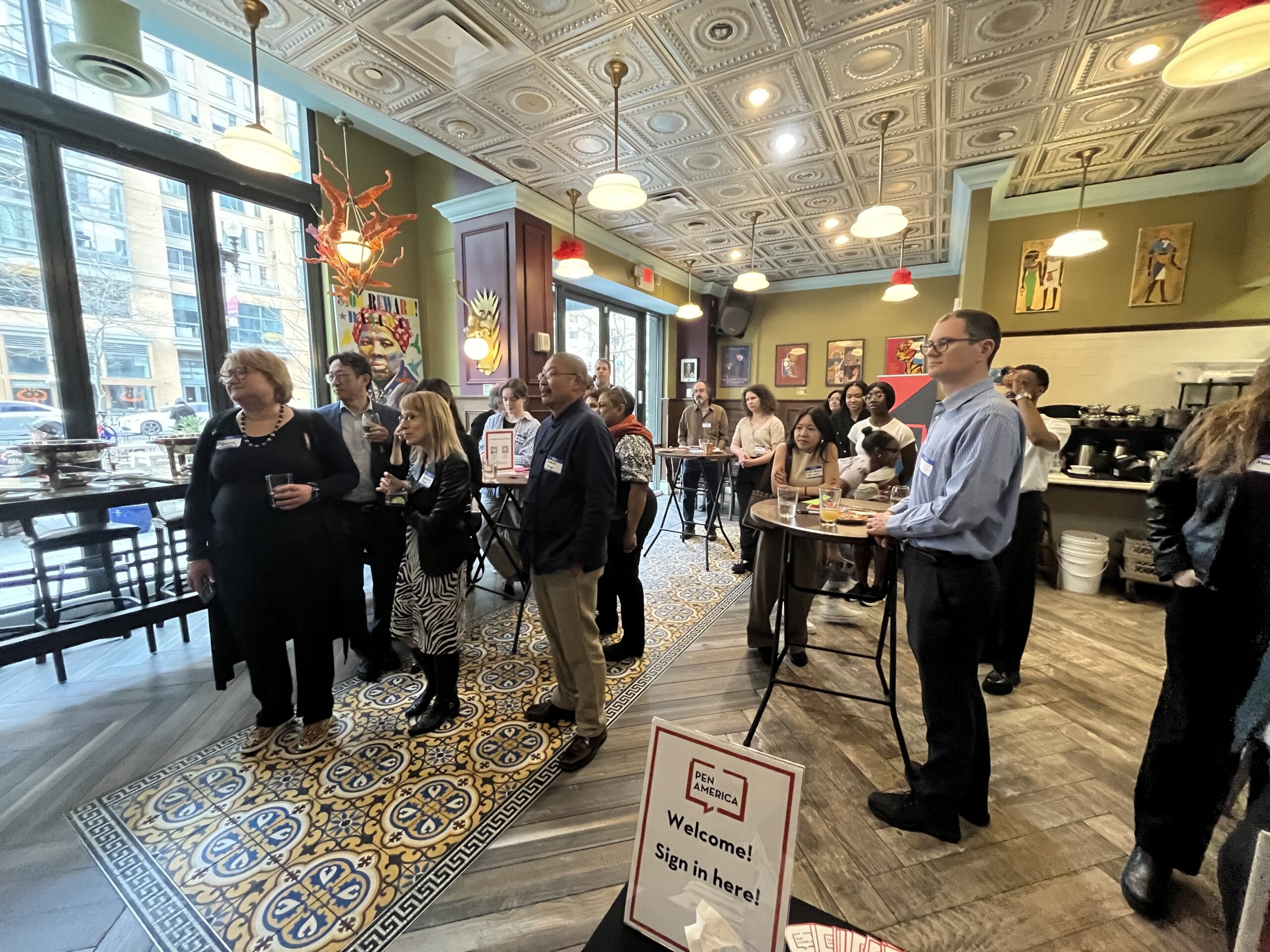 A group of people stand in a room with colorful artwork and a sign that reads Welcome! Sign in here! Some are seated while others stand at round tables, attentively listening. Large windows let natural light in, illuminating the patterned floor tiles.