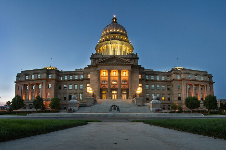 A majestic, illuminated capitol building with a prominent dome stands at dusk. The structure features columns and steps leading up to the entrance, with trees flanking both sides, under a clear, darkening sky.
