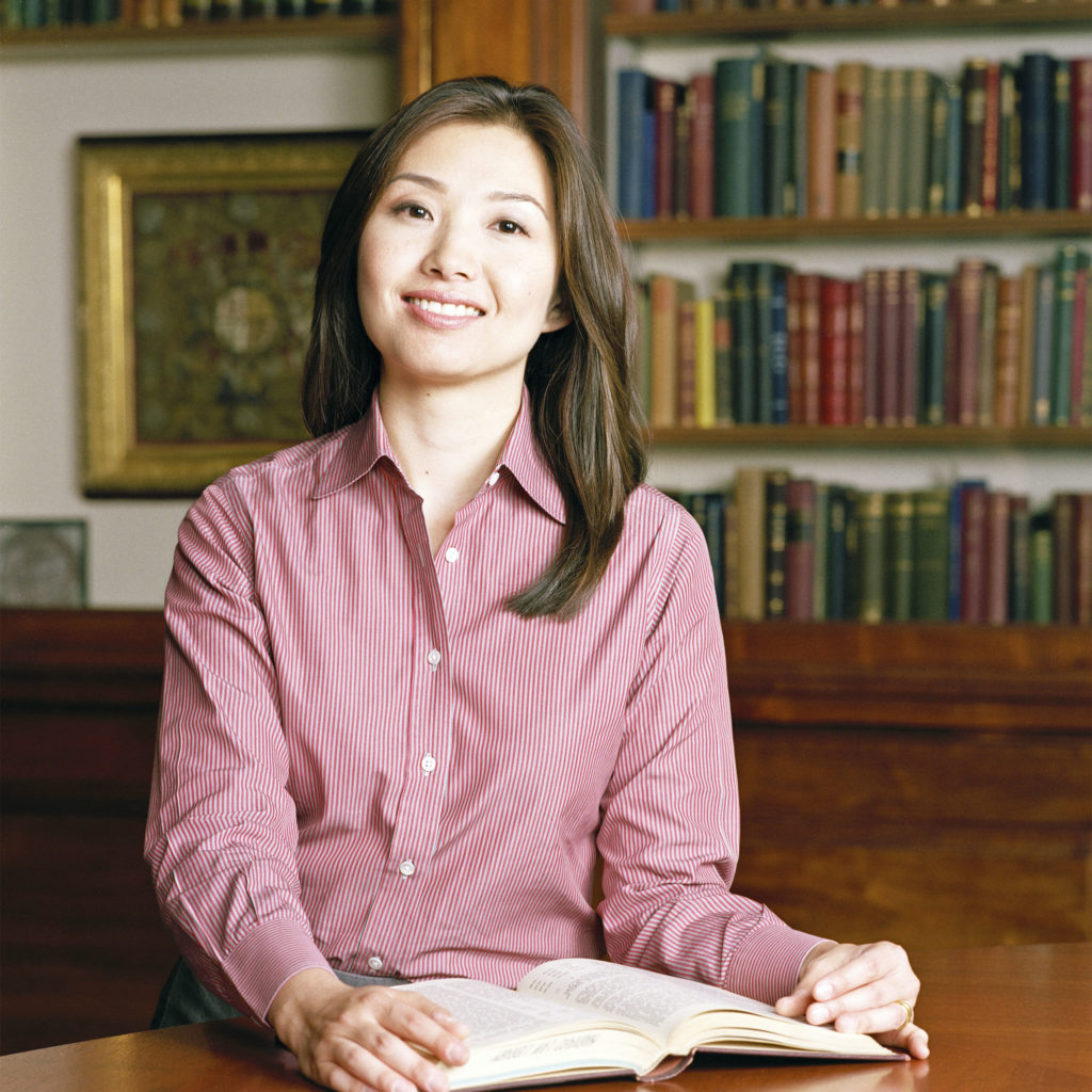 A woman with long brown hair, wearing a pink striped shirt, sits at a wooden table with an open book in front of her, smiling. Bookshelves filled with colorful books are in the background.