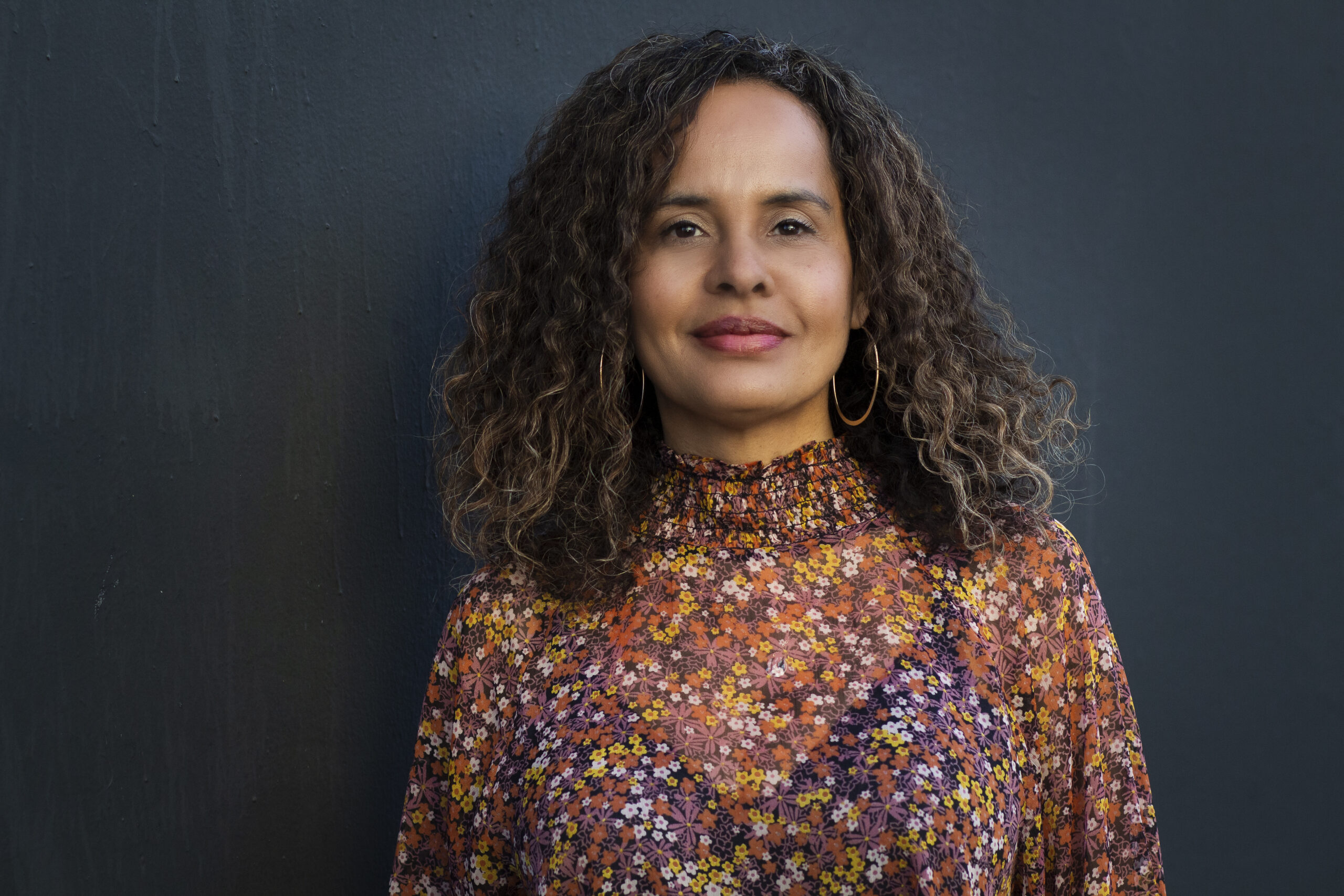A person with curly hair stands against a dark background, wearing a floral-patterned blouse with shades of orange and pink. They have a neutral expression and are looking directly at the camera.