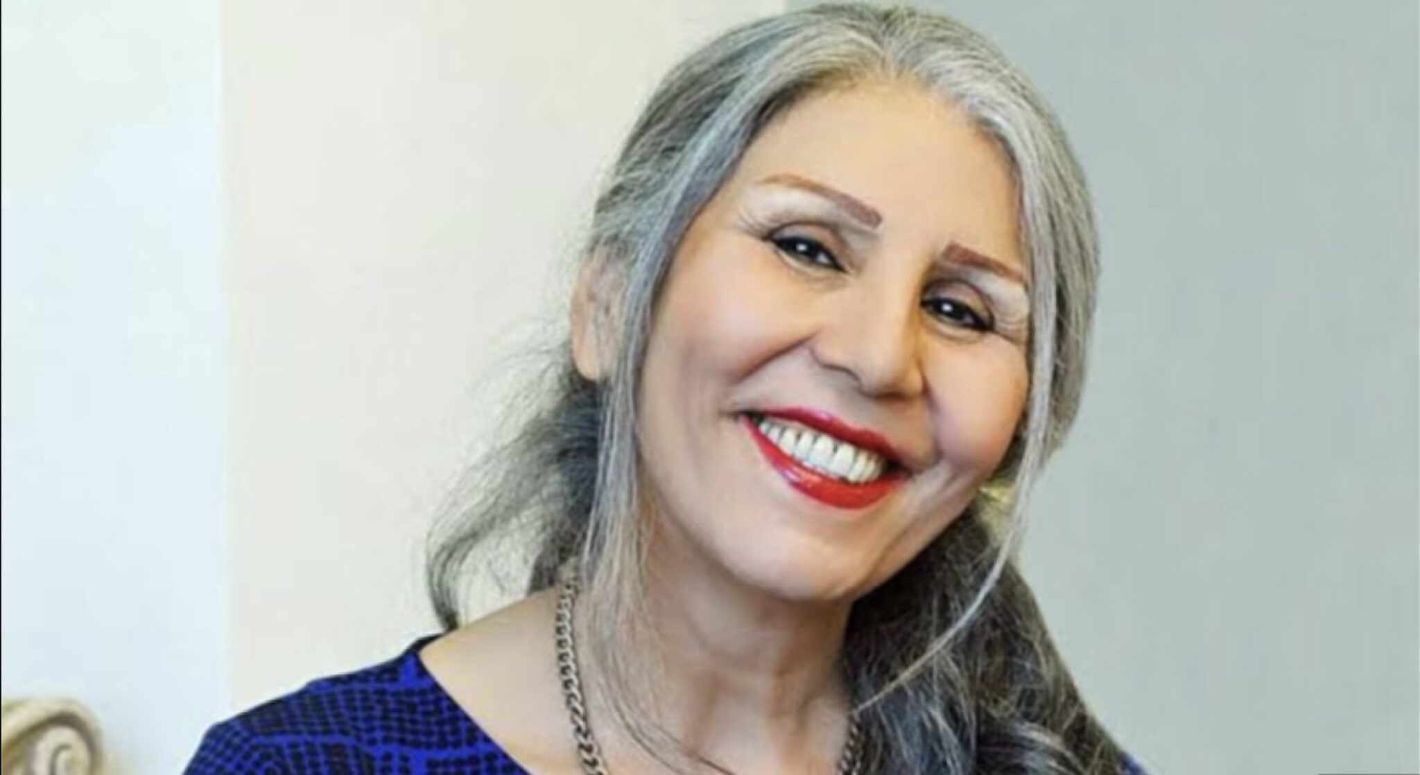 A woman with long gray hair, wearing a blue patterned top and a necklace, smiles warmly at the camera against a light background.