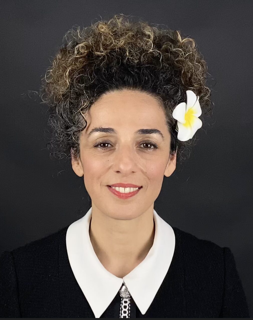 A woman with curly hair adorned with a white flower, wearing a black outfit with a white collar, smiles softly against a plain black background.