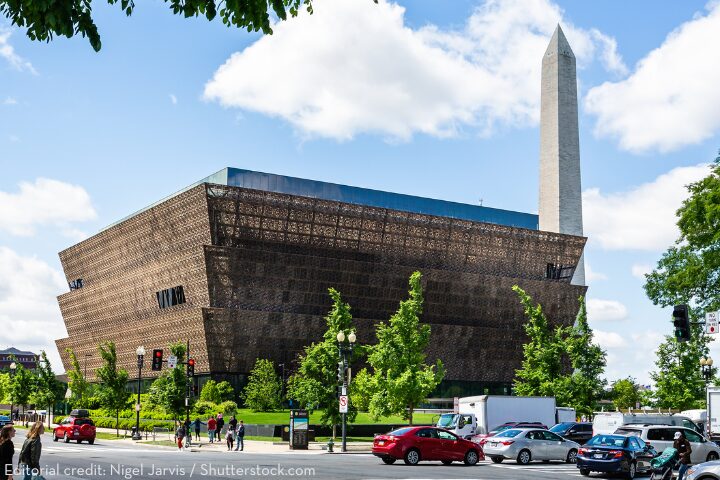 The National Museum of African American History and Culture stands near the Washington Monument in Washington, D.C., with trees, people, and cars visible in the foreground under a blue sky with clouds.