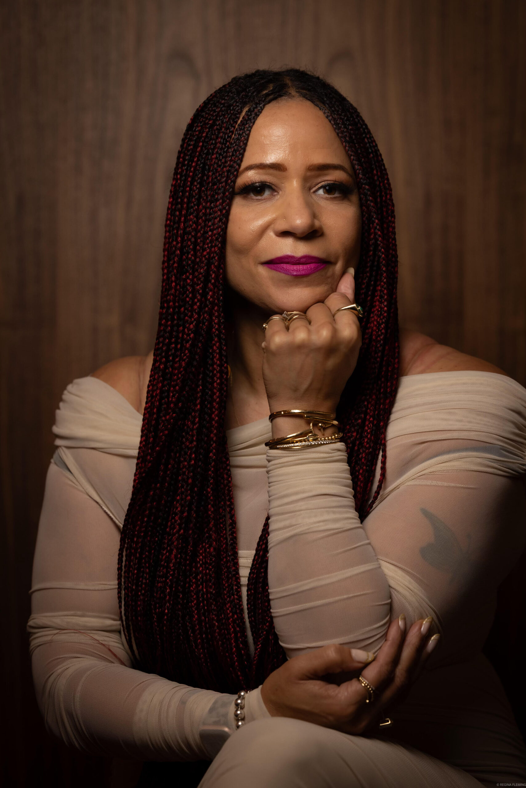 A woman with long, braided hair and wearing a sheer, off-the-shoulder beige top sits against a wooden background, resting her chin on her hand and looking confidently at the camera.