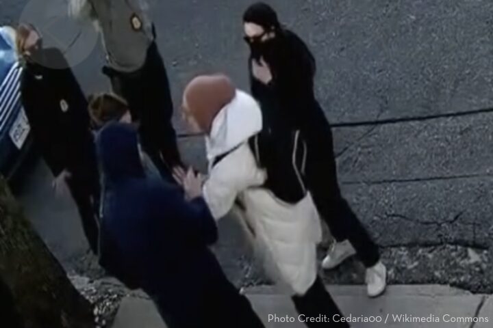 A group of four people, most wearing dark clothing and face coverings, surround a person in a white coat and brown hat on a city sidewalk near a parked car. One person appears to be gesturing emphatically.