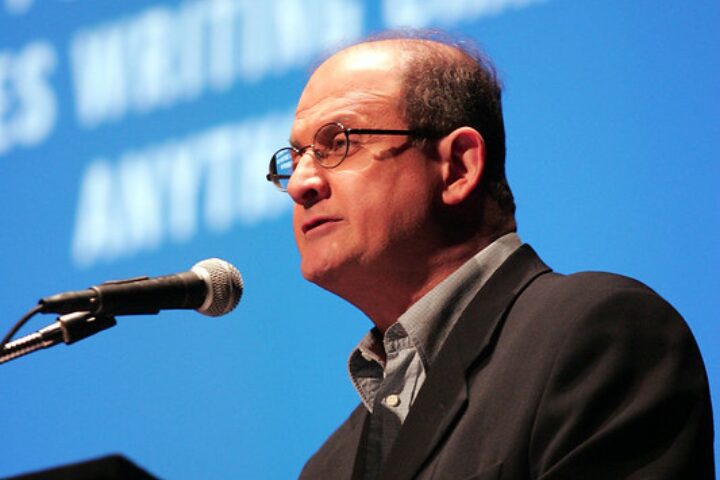 A man in glasses and a suit speaks into a microphone against a blue background with blurred white text, celebrating the World Voices Festival 20th Anniversary.