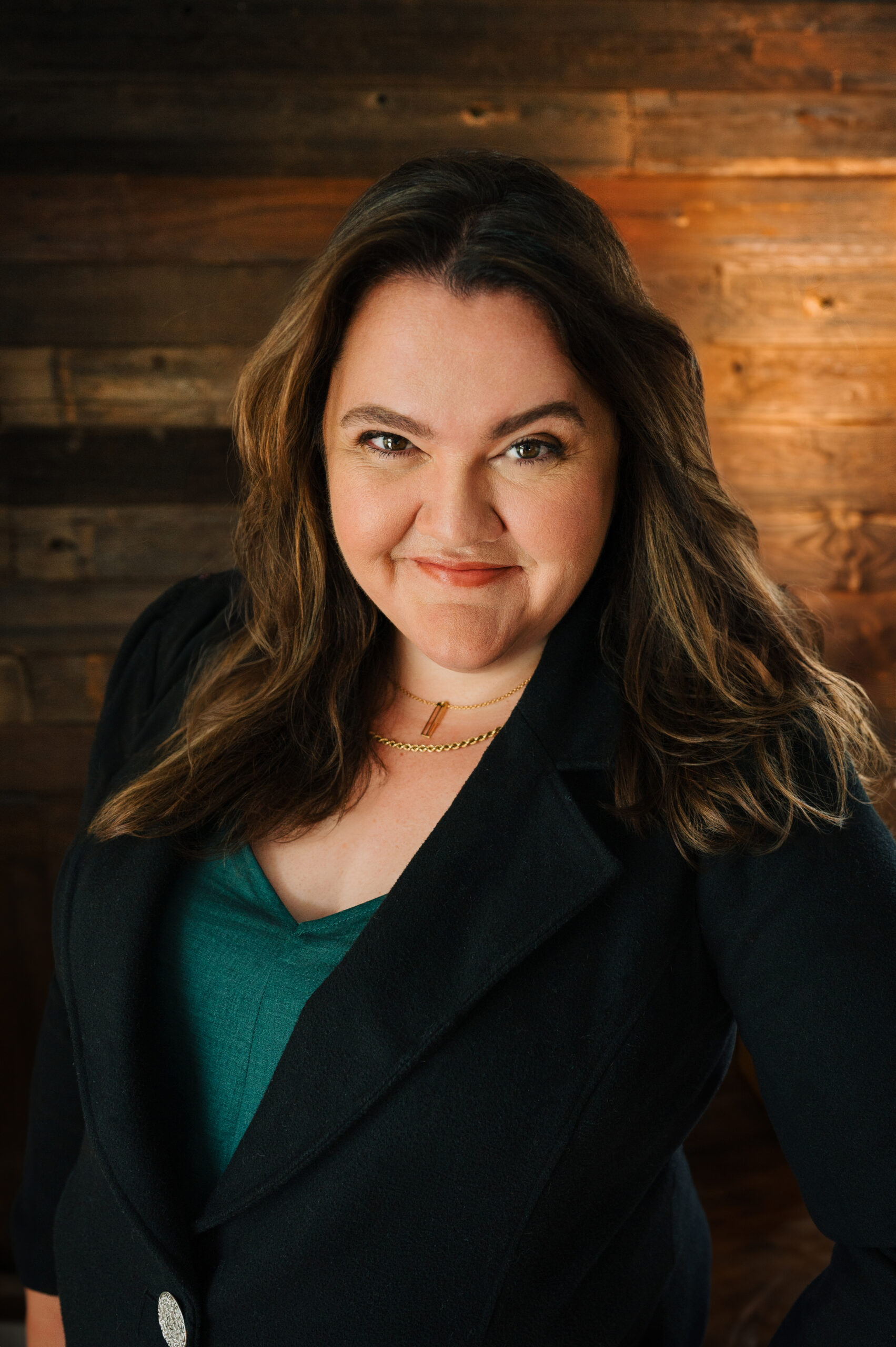 A woman with long brown hair wearing a black blazer and green top smiles confidently at the camera, standing against a wooden plank background.