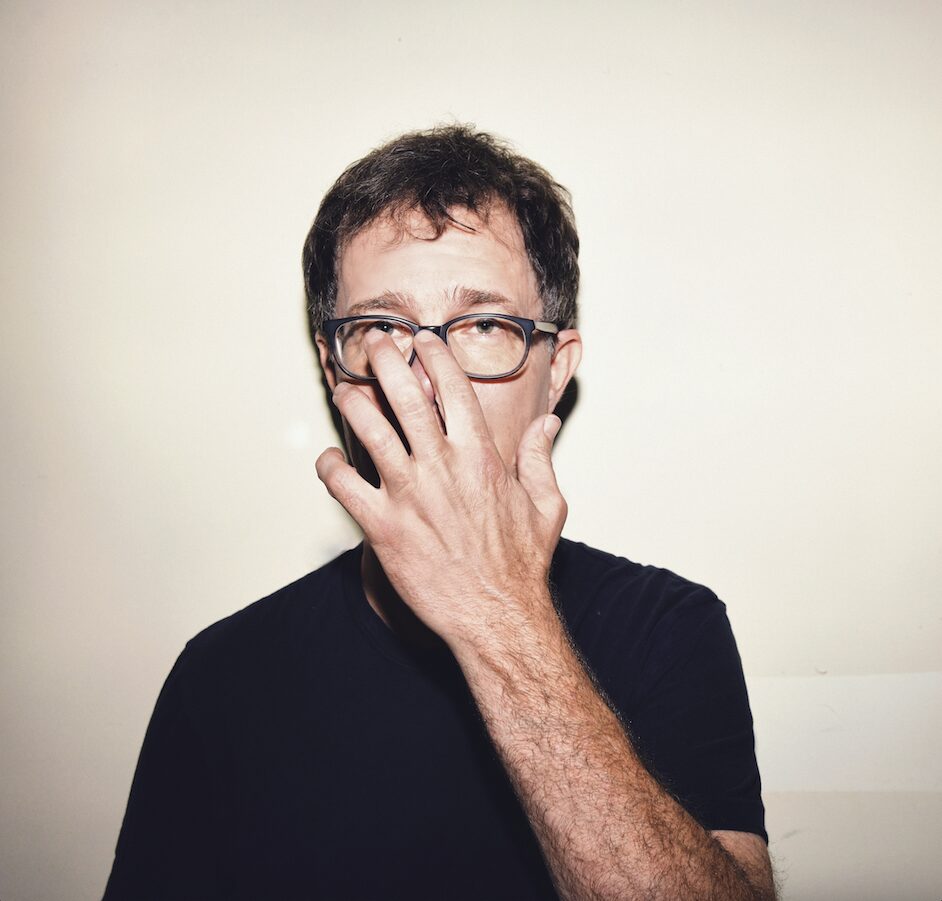 A man wearing glasses and a dark shirt stands against a plain light-colored wall, covering the lower half of his face with his hand.