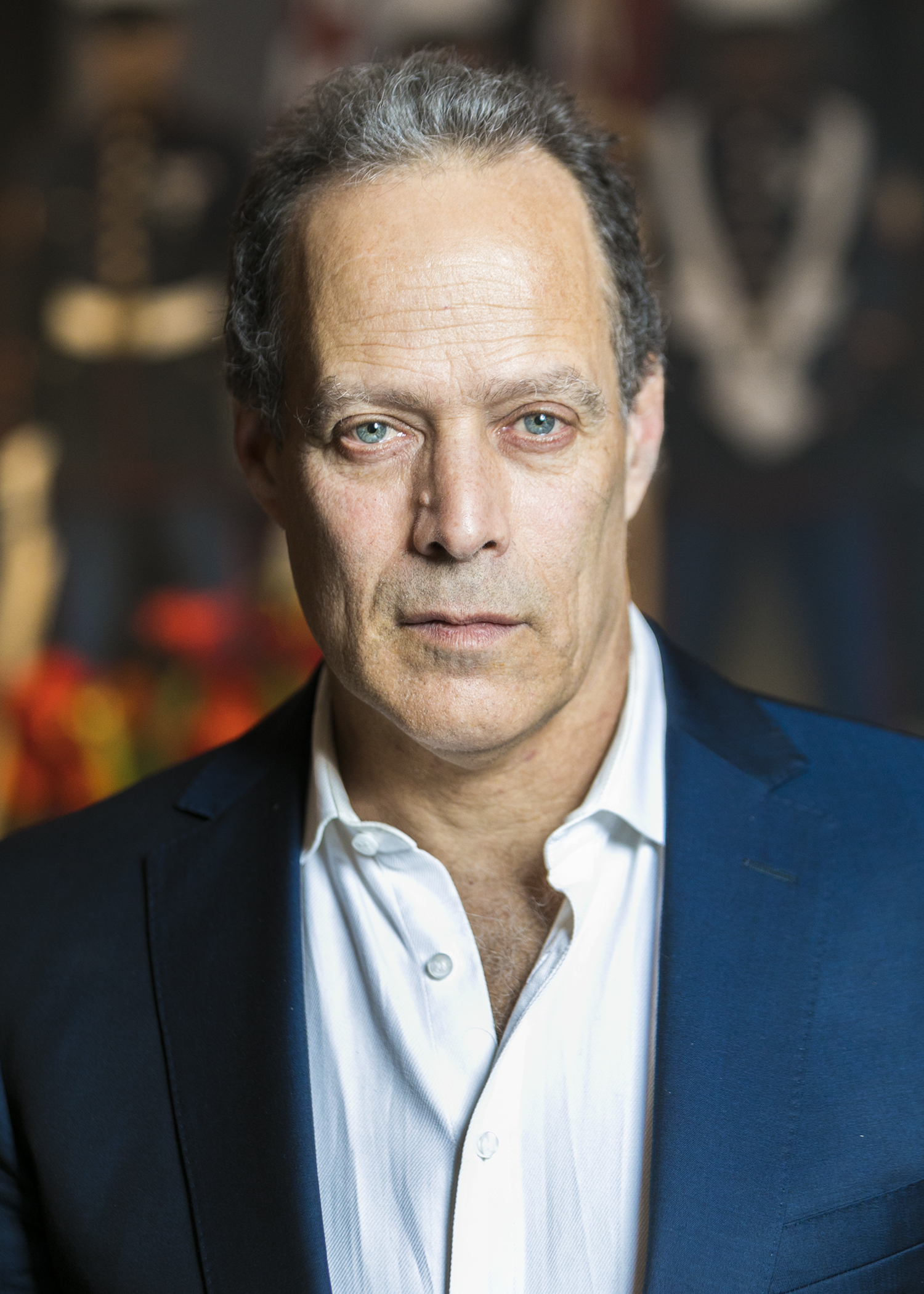 A middle-aged man with short curly hair wearing a white dress shirt and navy blazer, standing in front of a blurred background featuring colorful, out-of-focus figures.