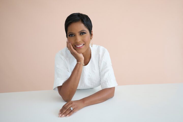 A woman with short hair smiles softly, leaning on a white table with her chin resting on her hand. She is wearing a white top, hoop earrings, and a ring, set against a light peach background.