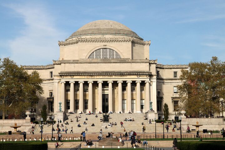 A large neoclassical building with a domed roof and tall columns, surrounded by trees. Many people are sitting or walking on the wide steps and plaza in front of the building under a clear sky.